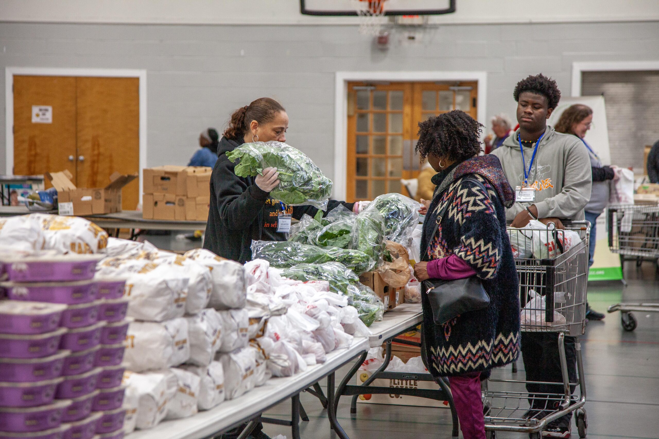 Volunteers distributing fresh produce and groceries at a community food pantry, with individuals selecting items from tables filled with packaged food and vegetables.