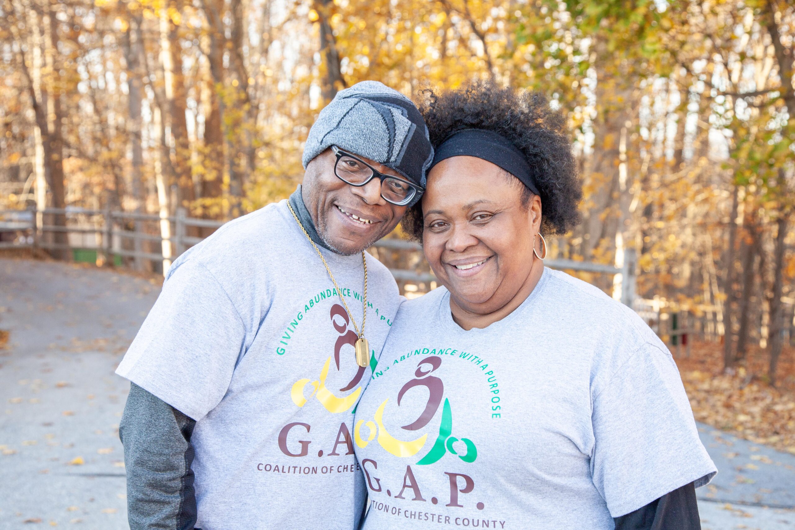 Smiling couple wearing G.A.P. Coalition of Chester County t-shirts, standing outdoors with autumn foliage in the background, promoting community engagement and support initiatives.