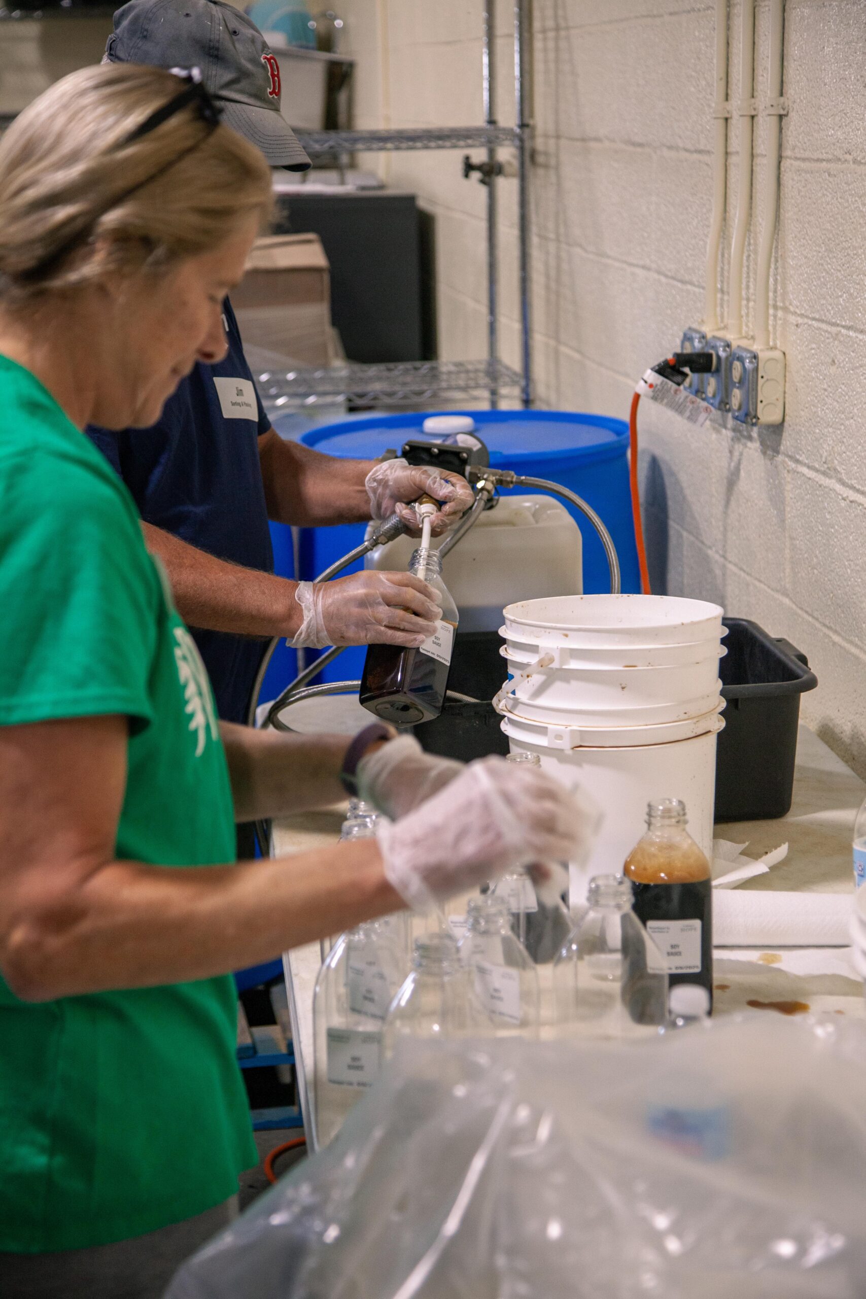 Two individuals in gloves are engaged in a laboratory setting, preparing and labeling various bottles with liquid samples. One person is pouring from a larger container into a smaller bottle, while the other is handling additional bottles on a table. The background features containers and equipment typical of a scientific workspace.