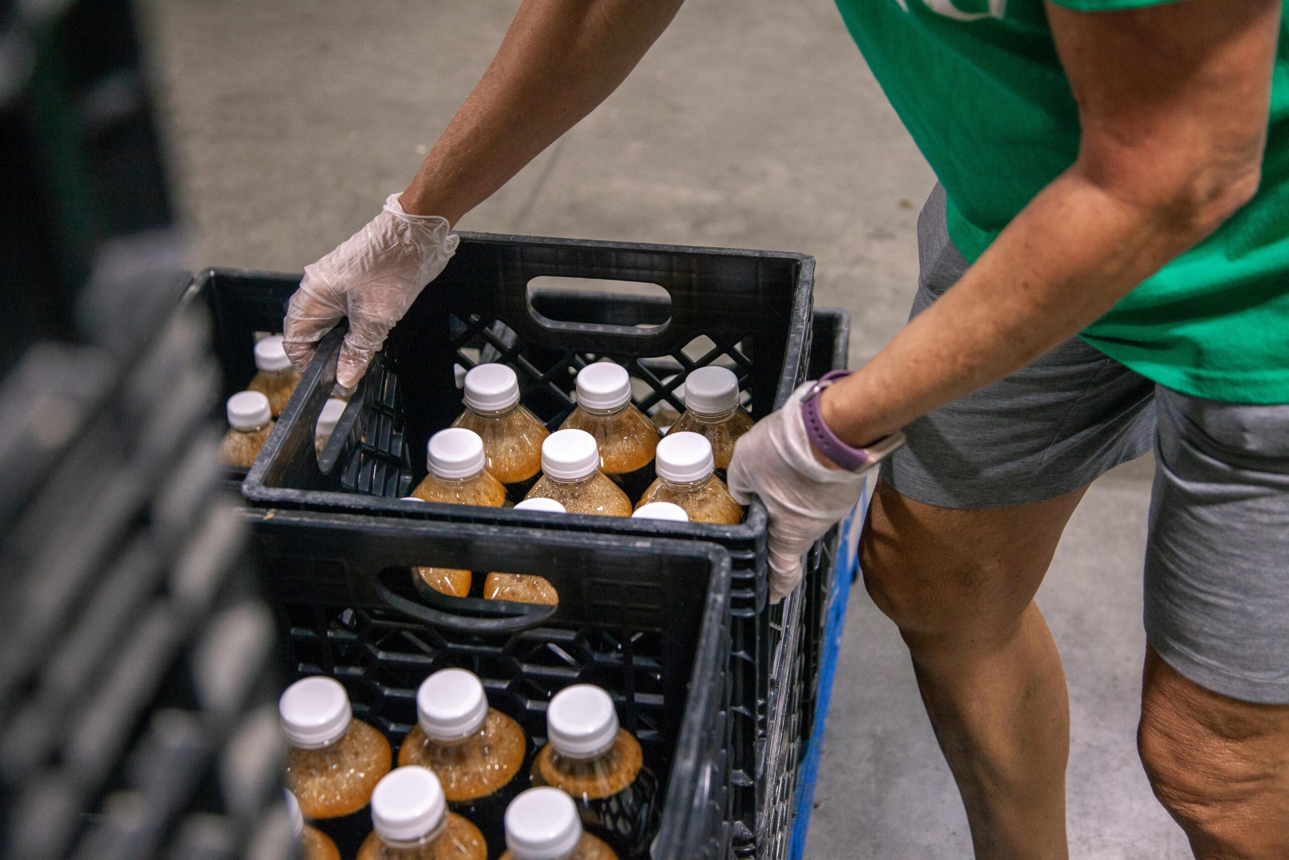 Person wearing gloves organizing crates of bottled beverages in a warehouse setting, emphasizing food safety and community support.