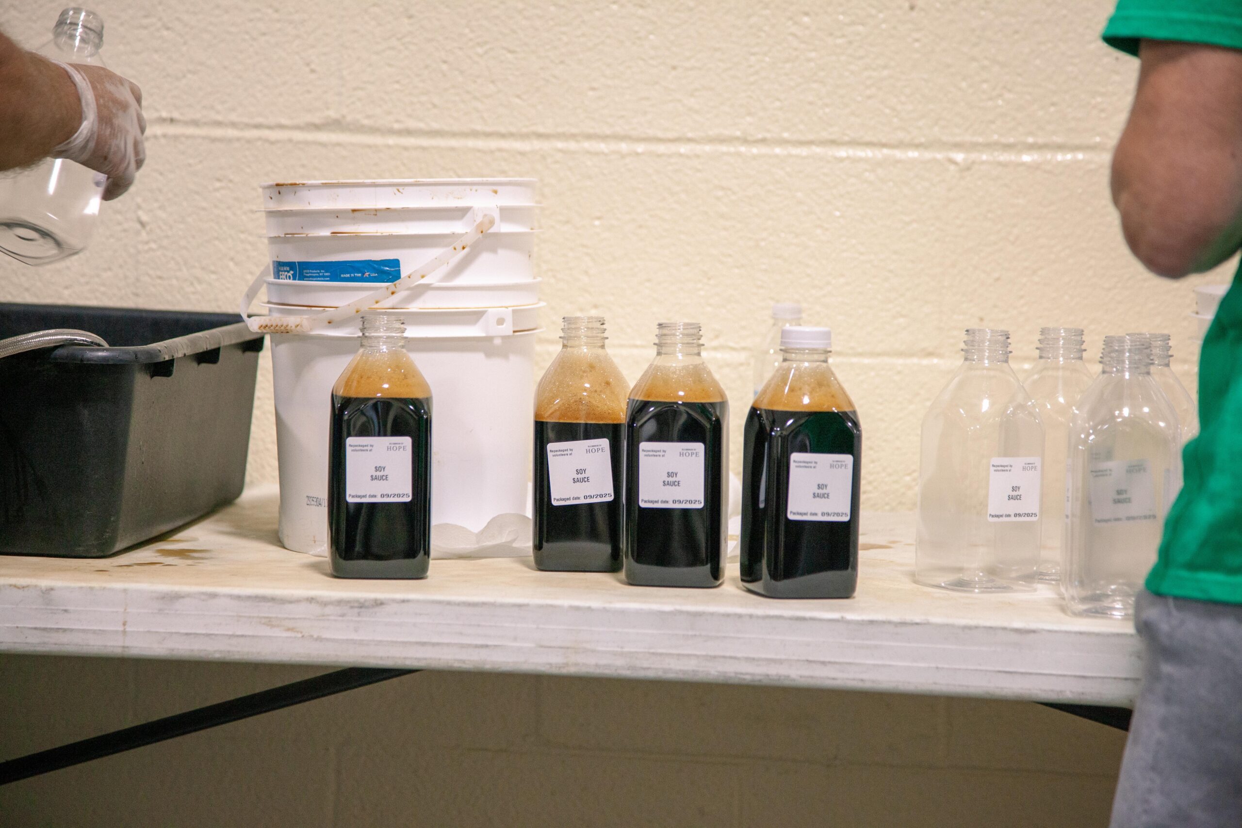 Bottles filled with soy sauce are lined up on a table, with one person's hand pouring from a glass container. A white bucket and a black bin are also visible in the background, indicating a food preparation or production setting.