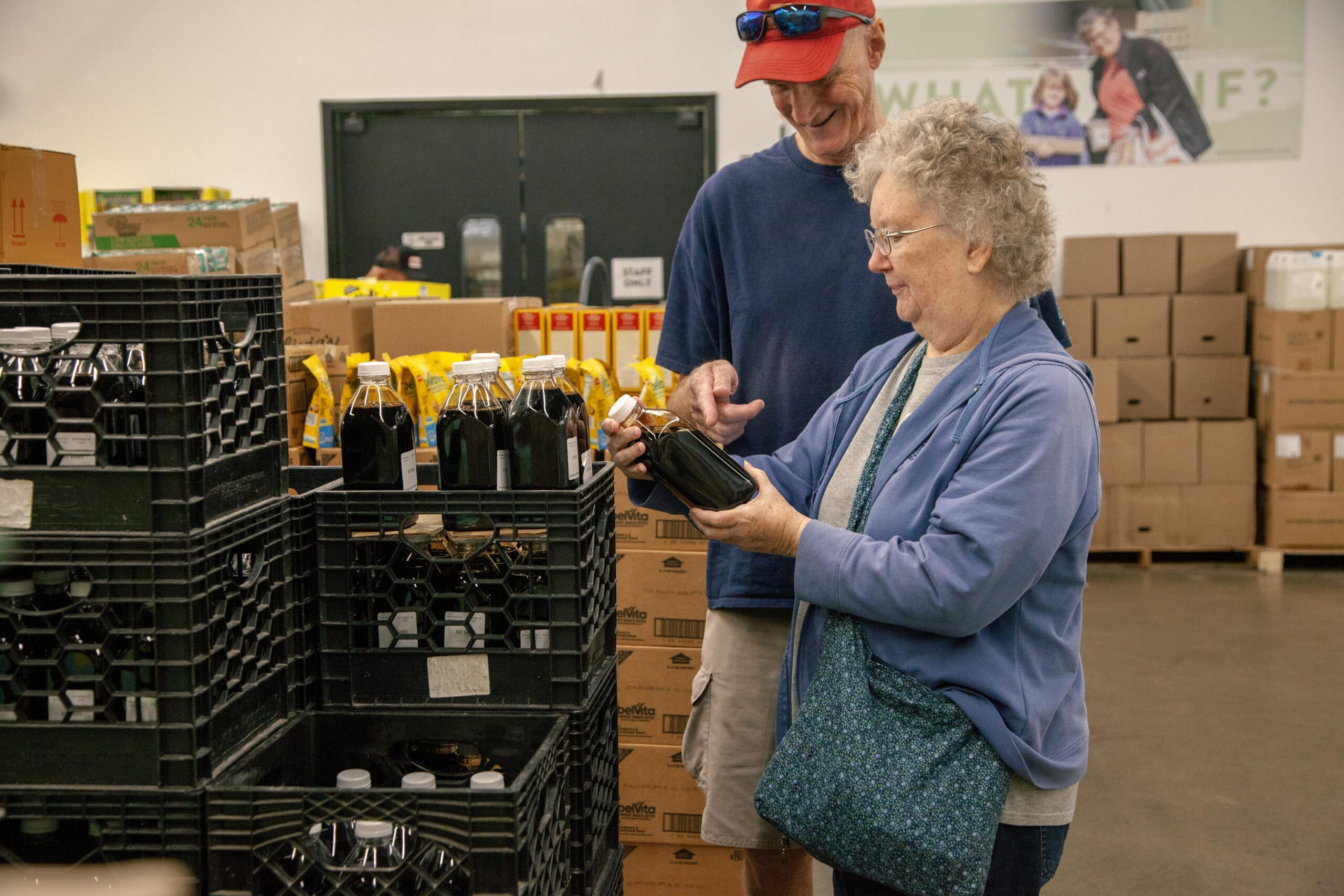 Elderly couple shopping at a grocery store, examining bottles of liquid in a crate, with shelves of products in the background.