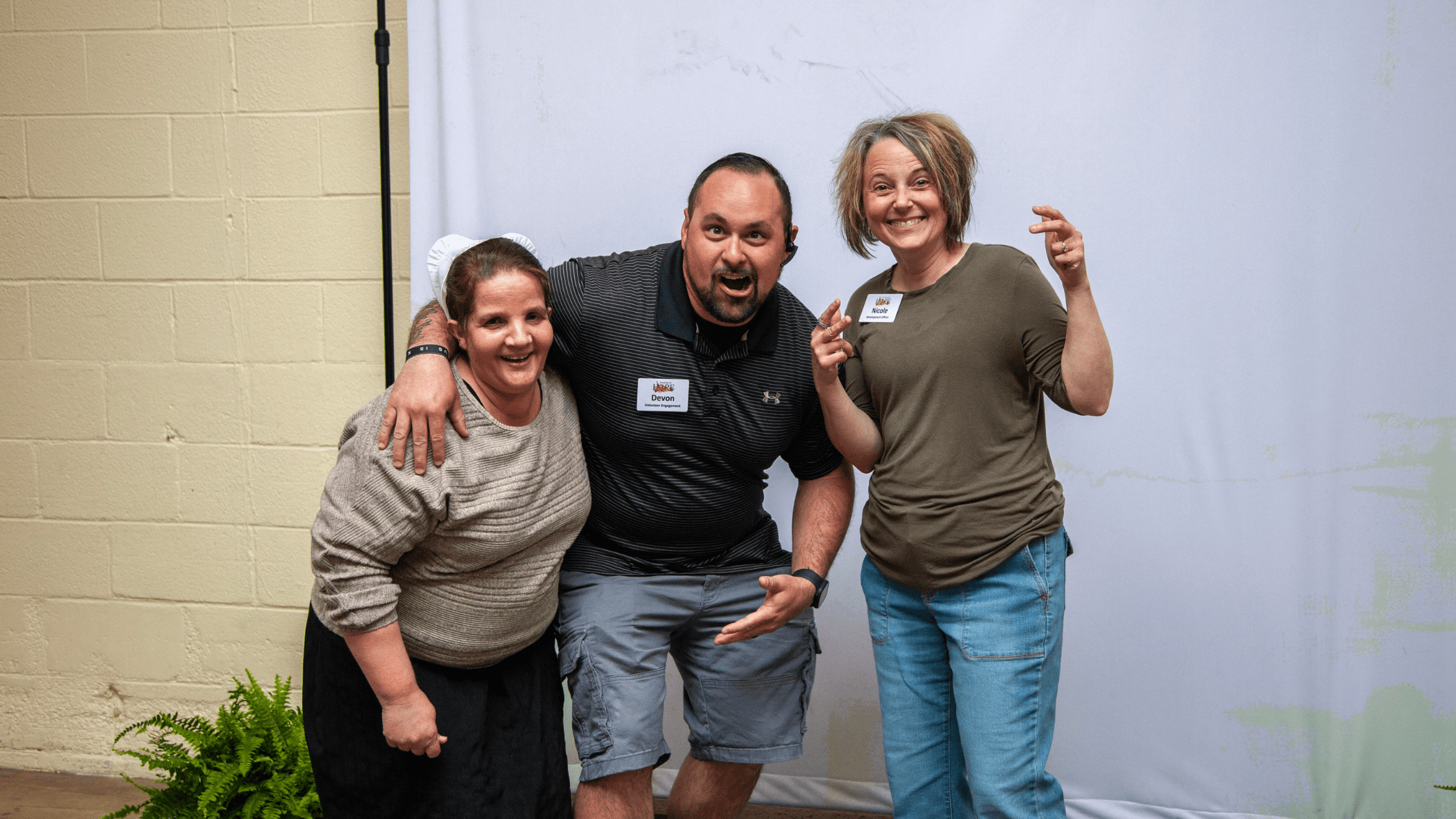 Three smiling individuals pose playfully in front of a blank backdrop, showcasing a sense of camaraderie and joy. The scene captures a lighthearted moment, ideal for representing friendship or community events.