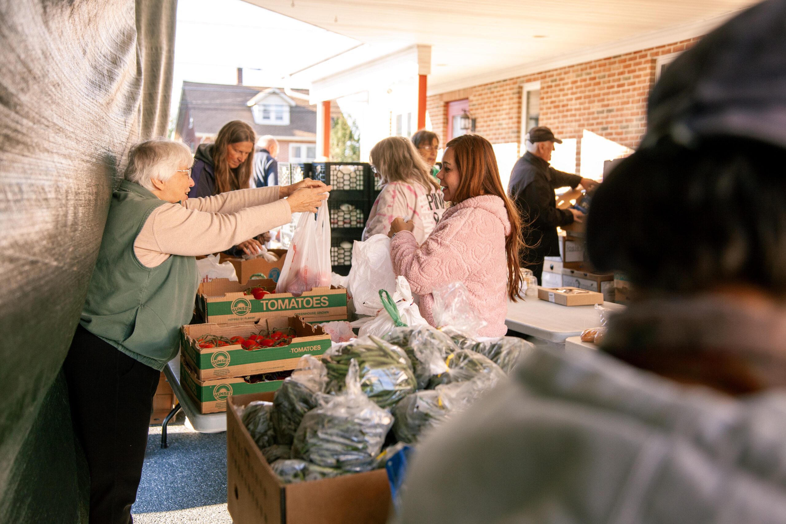 Community members gather at a local food distribution event, exchanging fresh produce such as tomatoes and green beans. The scene highlights the importance of community support and access to healthy food options.