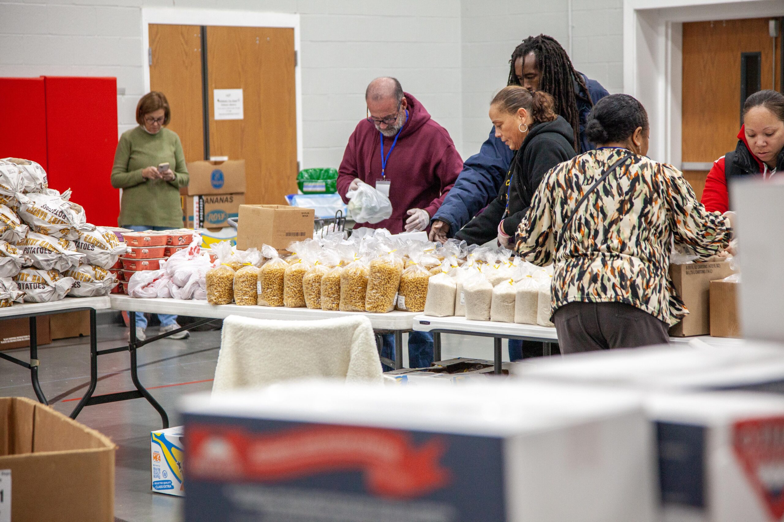 Volunteers sorting and packaging food items, including bags of rice and pasta, at a community food distribution event in a large indoor space.