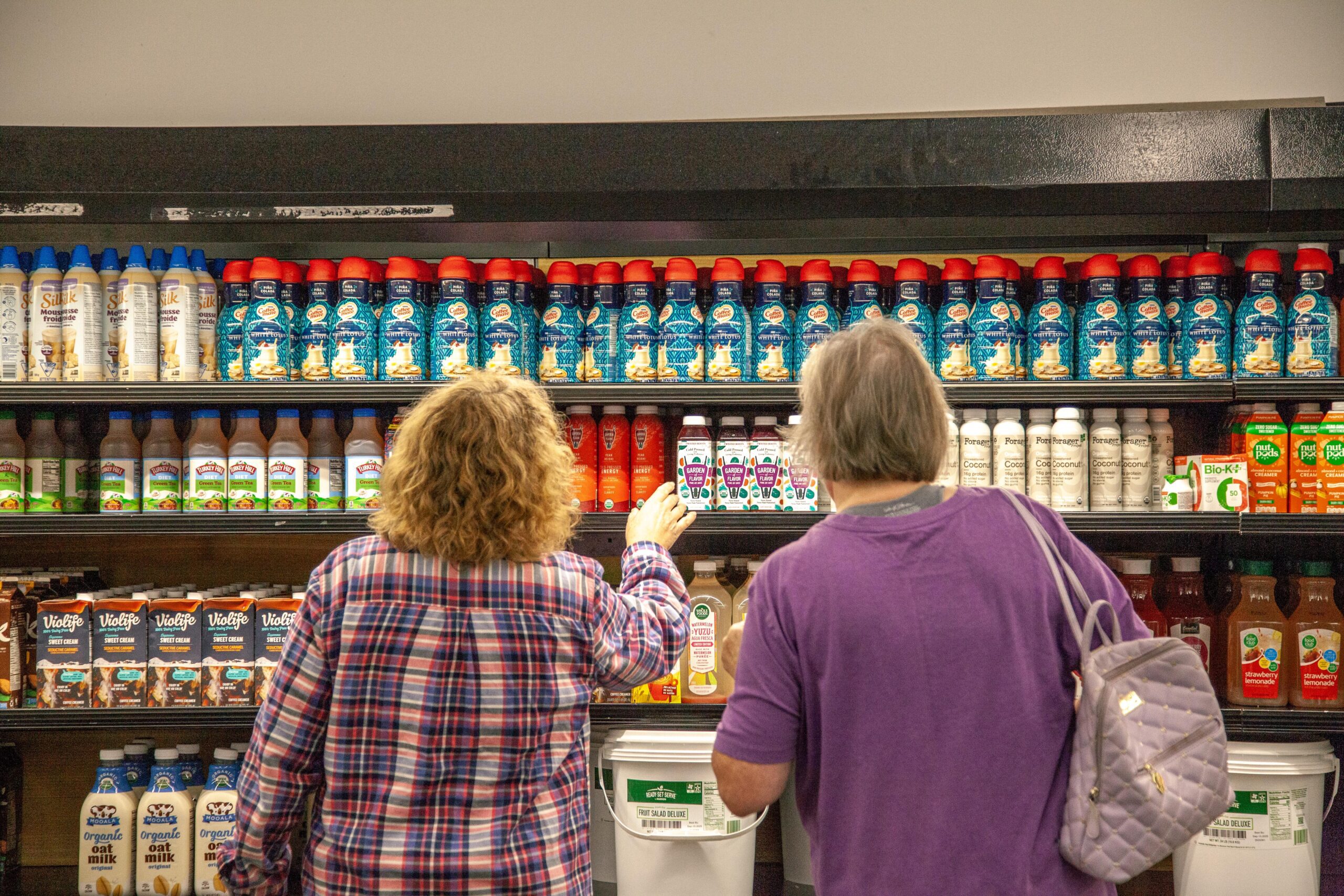 Two women shopping in a grocery store aisle filled with various flavored creamers, plant-based milk alternatives, and other beverages. The shelves display a colorful assortment of products, including oat milk, coconut cream, and chocolate milk.
