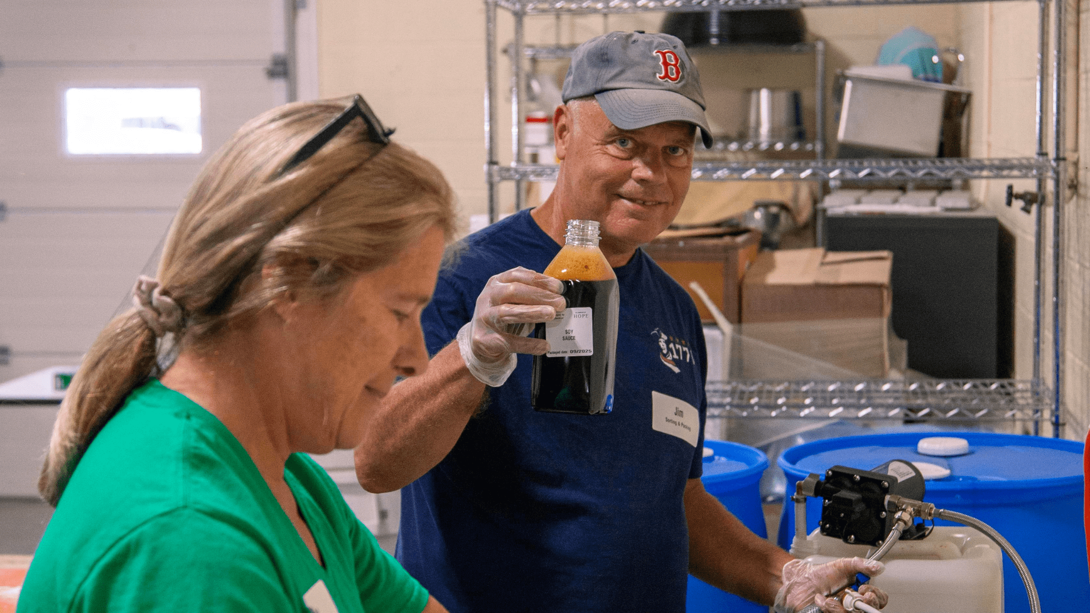 Volunteers preparing soy sauce in a kitchen setting, with a man in a baseball cap holding a bottle of sauce and a woman focused on her task, showcasing teamwork in food production.