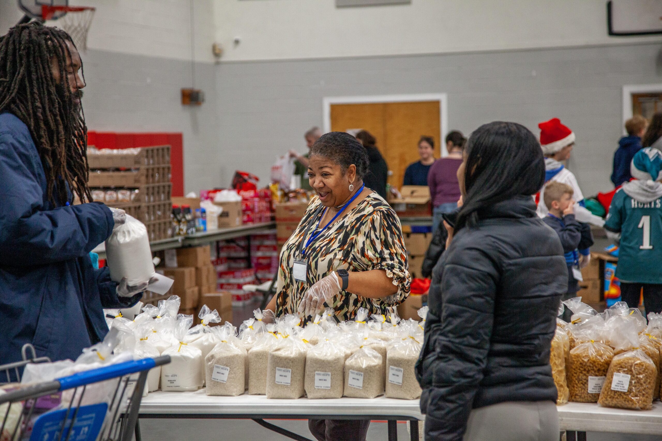 Volunteers distributing packaged food items at a community food pantry event, with a focus on a woman interacting with a man while surrounded by bags of rice and pasta on a table.