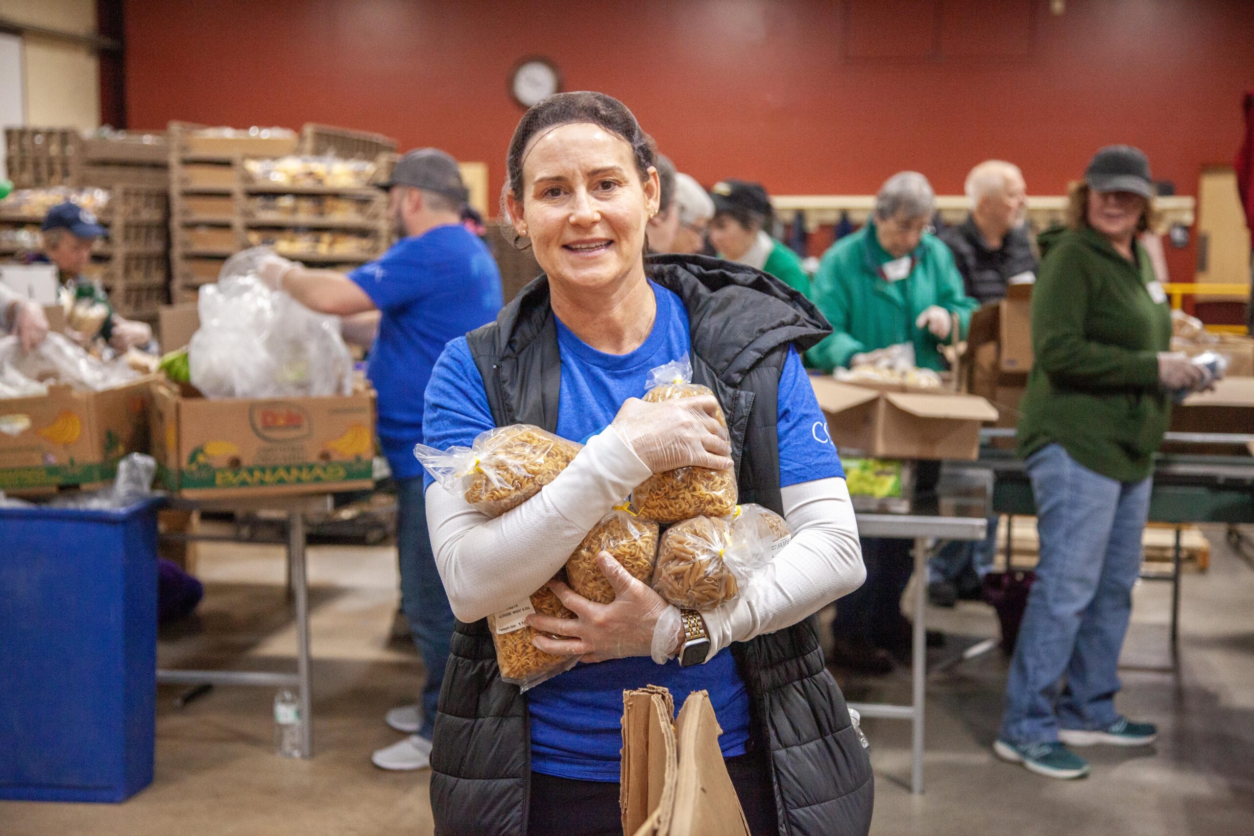 A volunteer at a food bank smiles while holding multiple bags of pasta, surrounded by others packing food in a community kitchen setting.