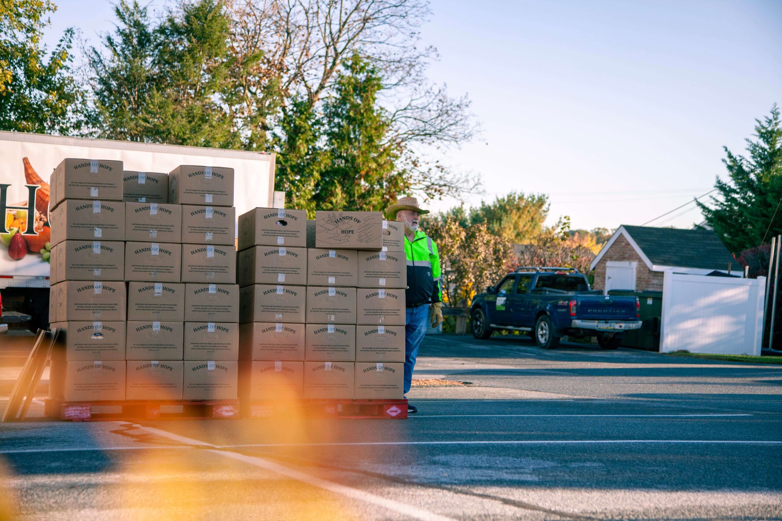 A worker in a reflective vest stands beside a stack of cardboard boxes labeled "Hands of Hope," with a blue pickup truck parked nearby and trees in the background, during daylight.