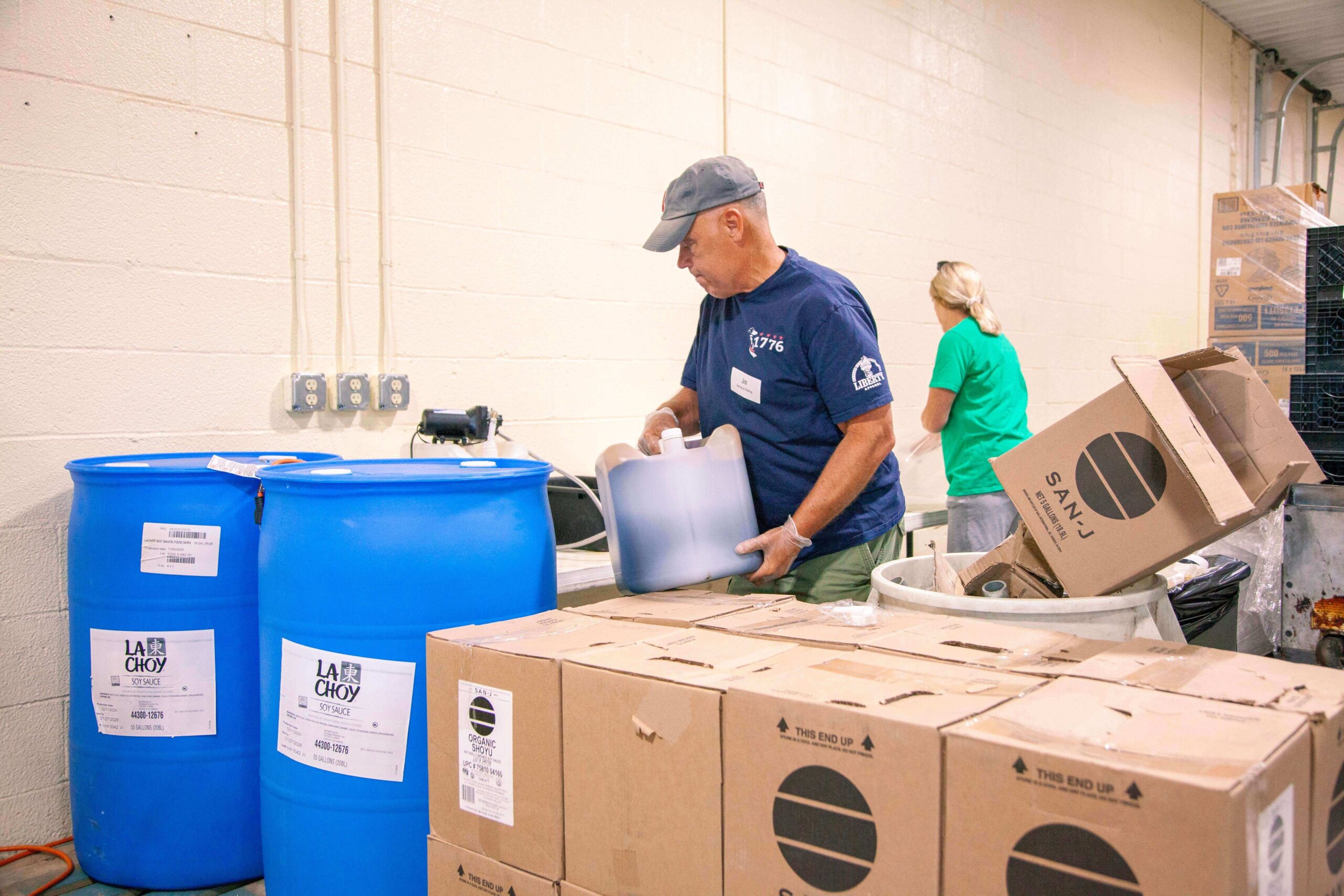 Volunteers working in a food distribution center, handling blue barrels of soy sauce and cardboard boxes, demonstrating community service and food preparation efforts.