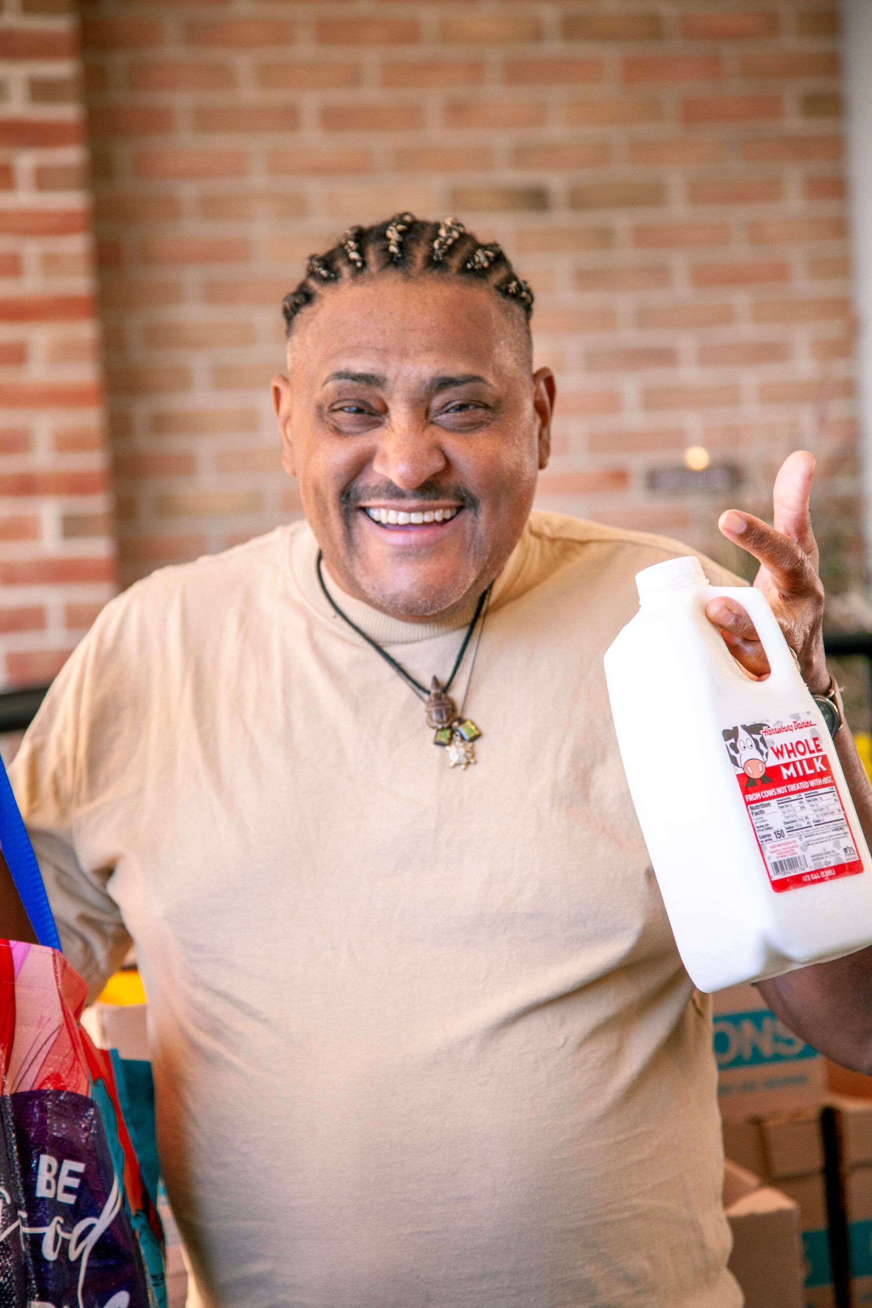 Smiling man holding a gallon of whole milk while standing in a kitchen with a brick wall and grocery bags nearby, promoting community wellness and healthy eating.