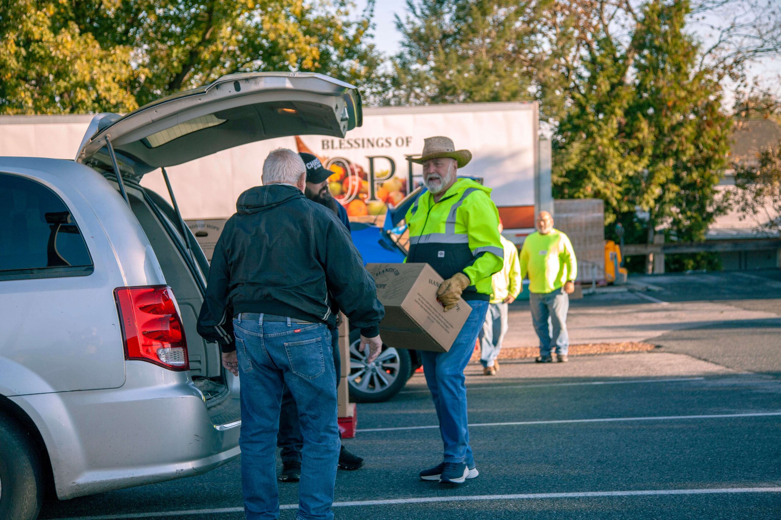 Volunteers unloading boxes from a minivan at a community food distribution event, with a large truck in the background displaying "Blessings of Hope." The scene captures the spirit of giving and community support.