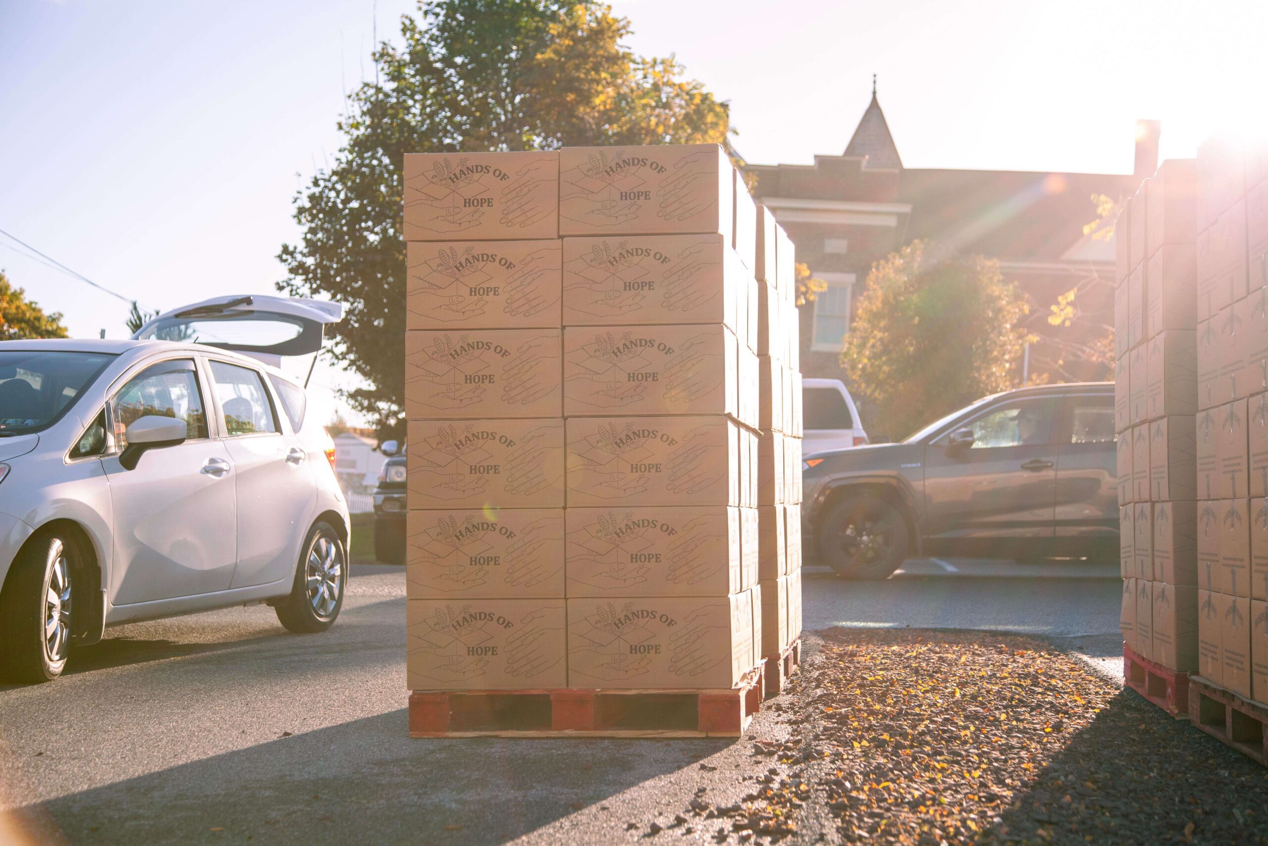 Pallets stacked with "Hands of Hope" boxes beside parked cars on a sunny street, highlighting community support and charitable efforts.