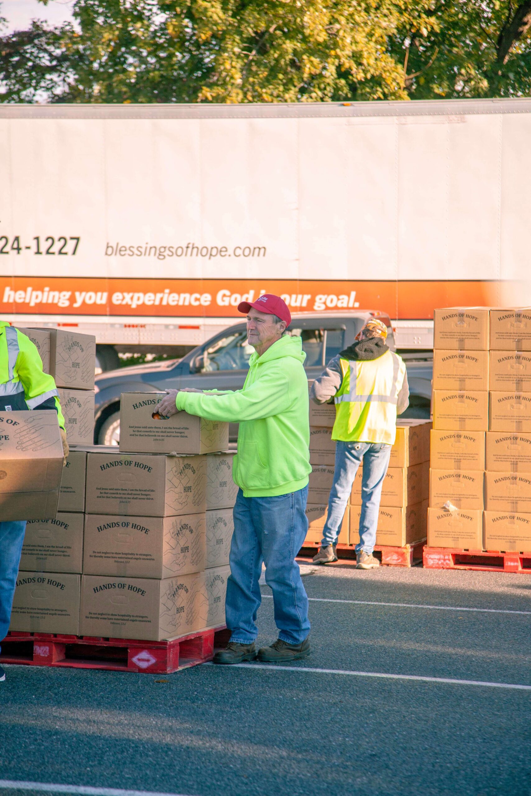 Volunteers unloading and distributing boxes labeled "Hands of Hope" at a community food drive, with a large truck in the background displaying the Blessings of Hope logo.