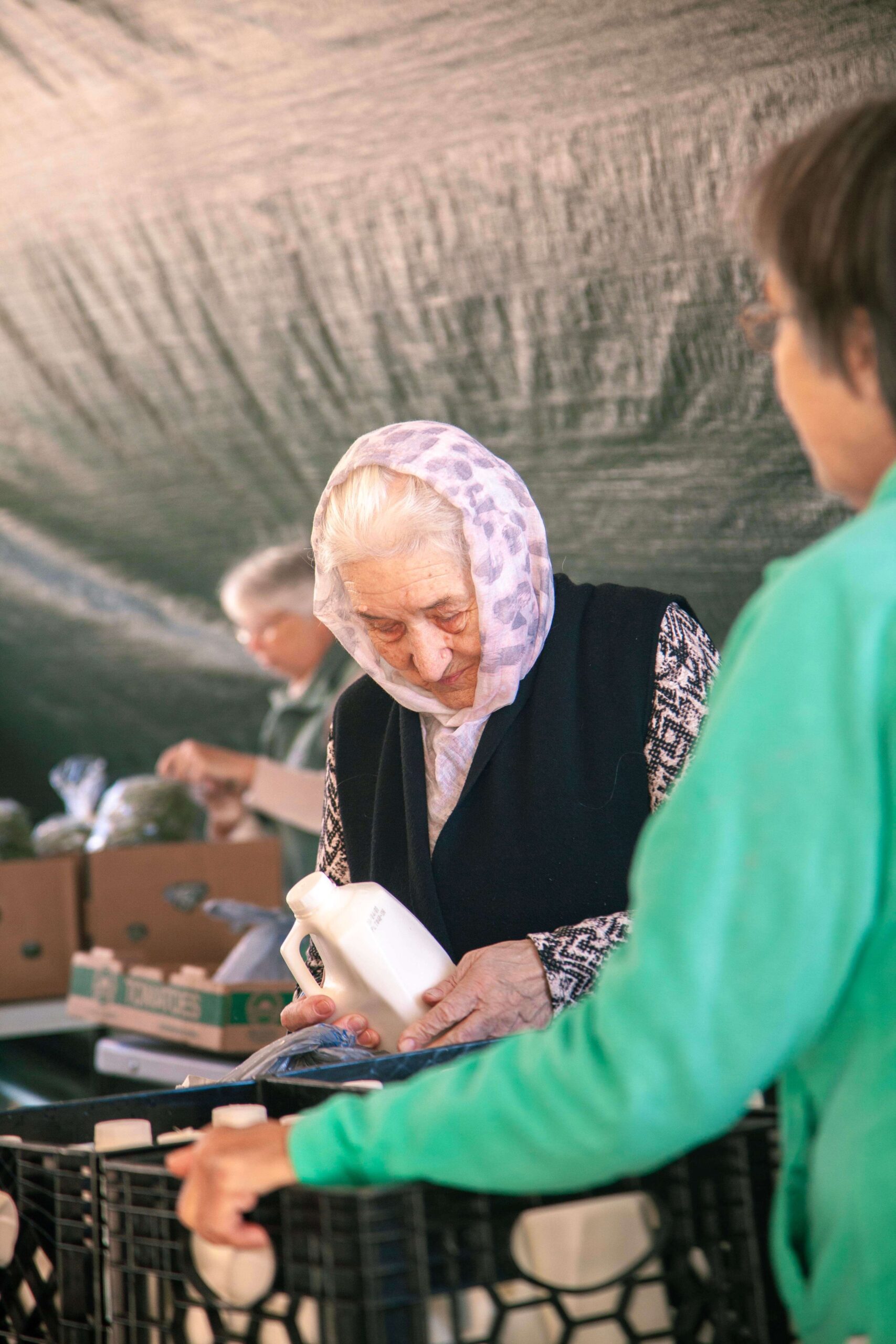Elderly woman wearing a headscarf examines a bottle while shopping at a market, with another person in a green jacket assisting her in the background. The scene captures the community spirit of local markets.