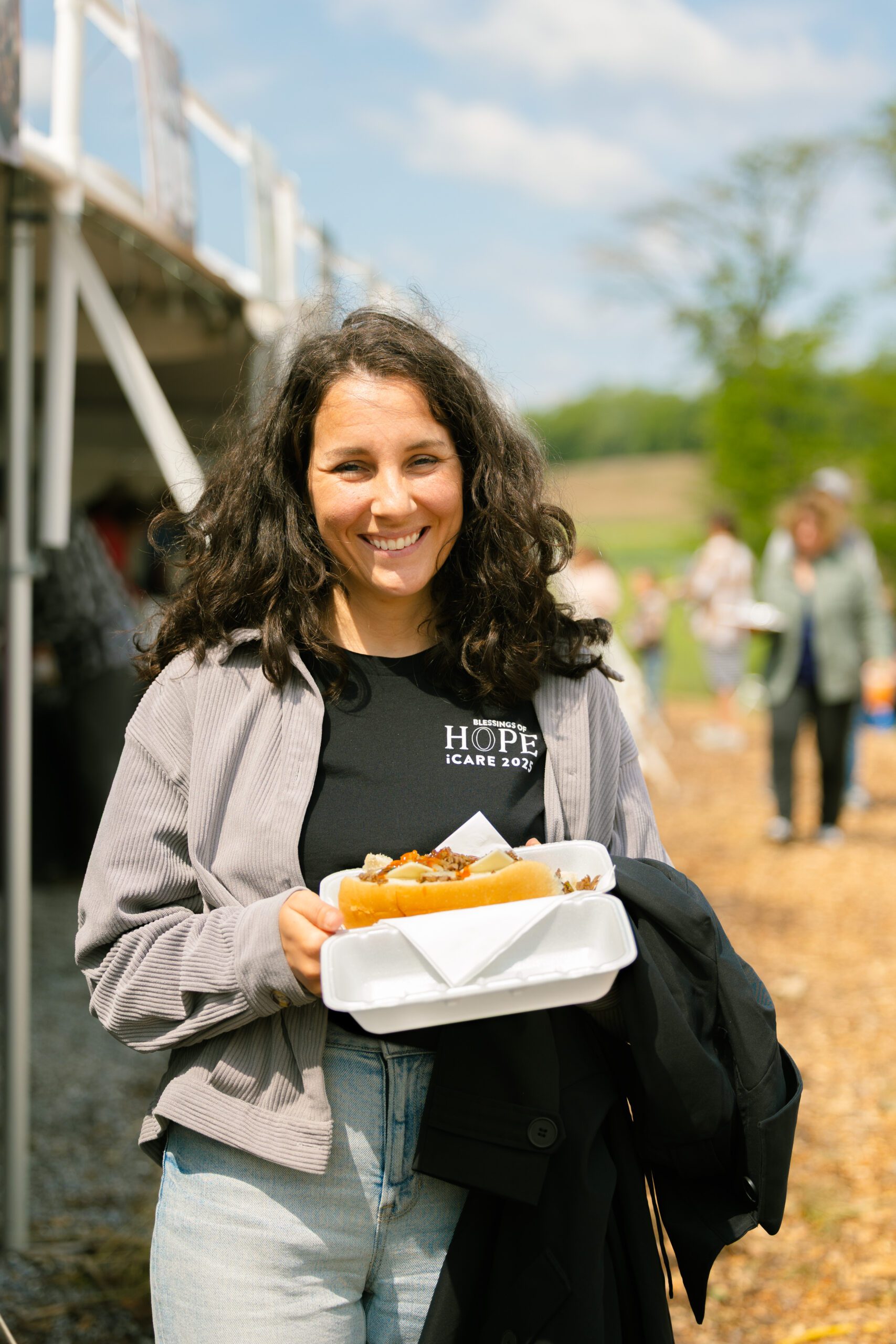 Smiling woman holding a takeout container with a sandwich at an outdoor event, wearing a black t-shirt with "Hope" and "iCARE 2023" printed on it, surrounded by people in a sunny, grassy setting.