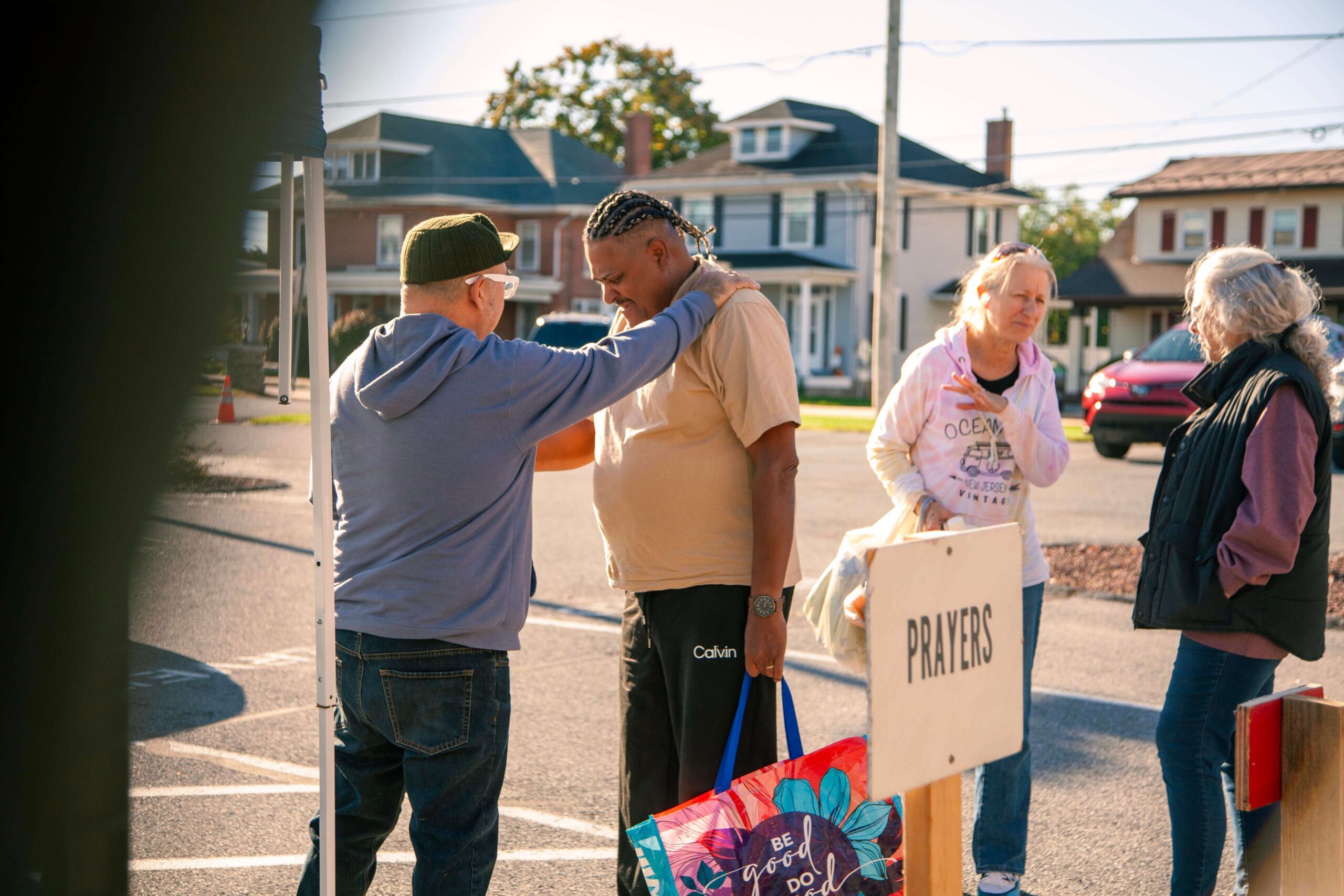 Community members engage in a prayer gathering, with one person comforting another in a heartfelt interaction. A sign labeled "PRAYERS" is visible, indicating the purpose of the gathering. The scene captures a warm, supportive atmosphere among individuals in a neighborhood setting.
