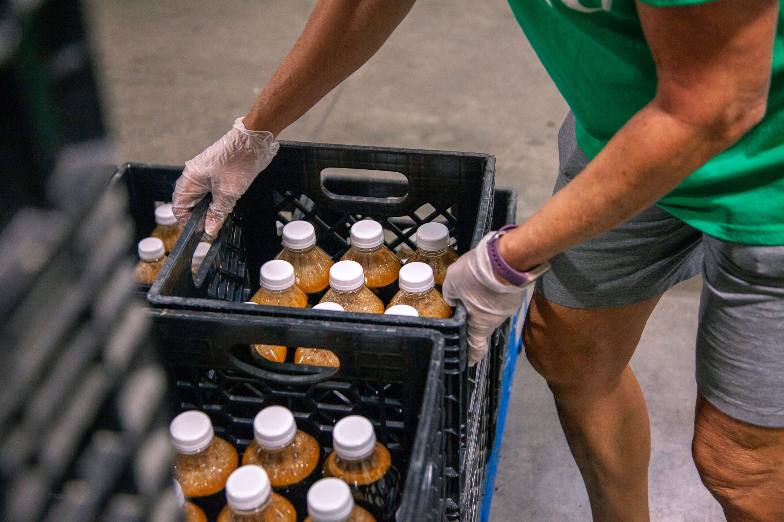 Person wearing gloves organizing crates filled with bottled beverages in a warehouse setting, highlighting food distribution efforts and community support.