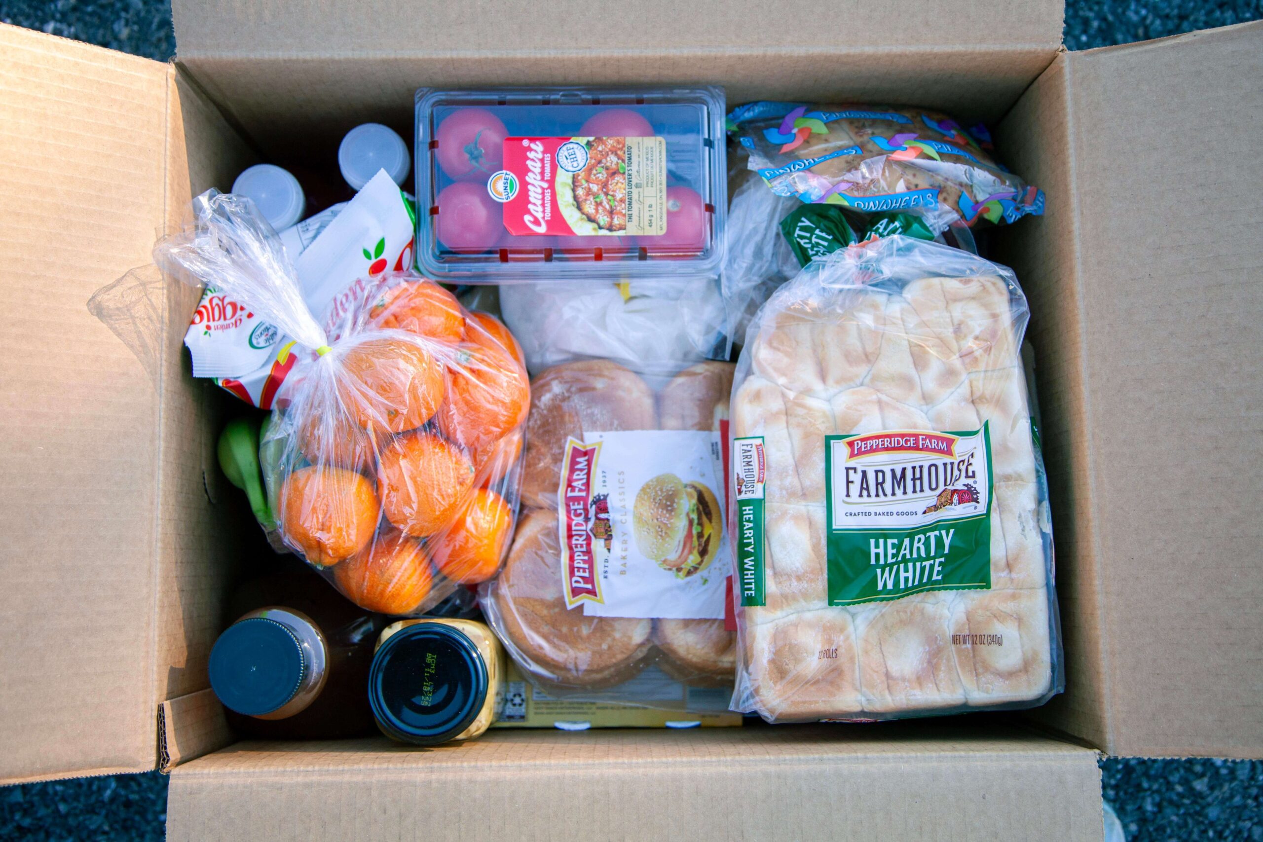 Cardboard box filled with various groceries including oranges, tomatoes, bread, and beverages, showcasing a selection of fresh produce and packaged food items.