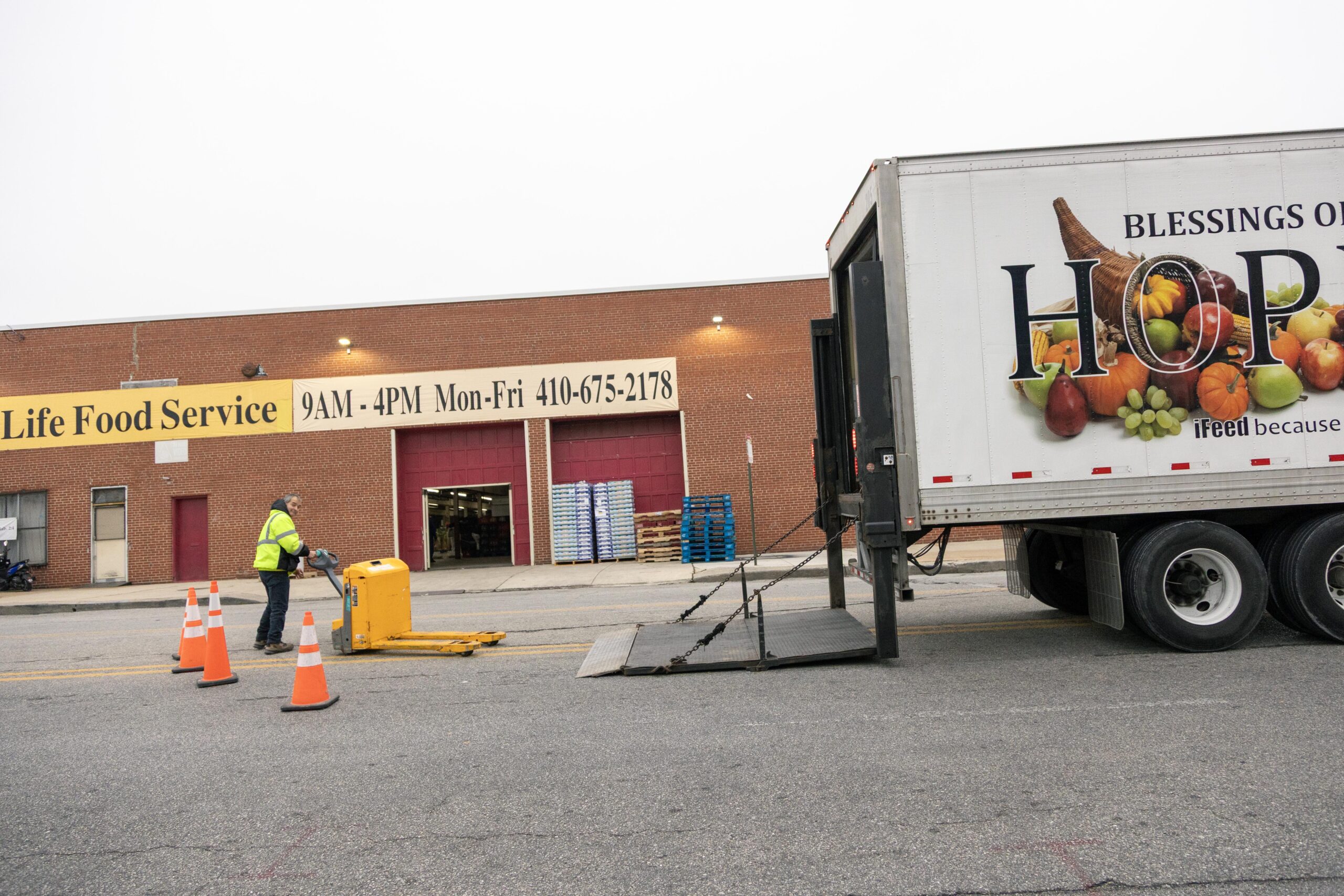 Worker unloading delivery at Life Food Service, a food distribution center, with visible signage showing operational hours and contact information. A truck featuring a colorful produce graphic is parked nearby, indicating fresh food supply efforts.