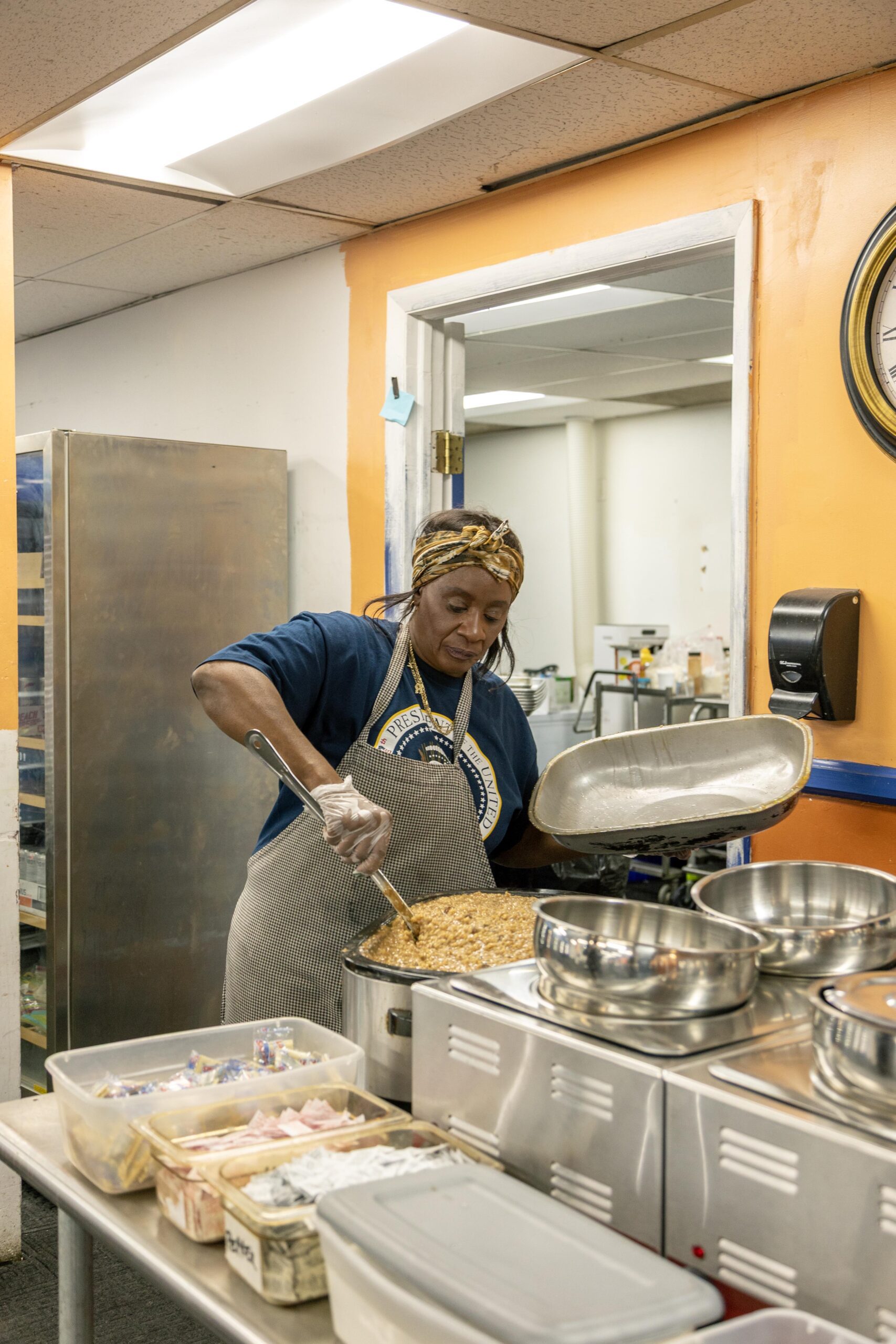 A woman in an apron prepares a large pot of food in a community kitchen, surrounded by cooking utensils and containers of ingredients. The kitchen features bright orange walls and stainless steel equipment, highlighting an active food preparation environment.