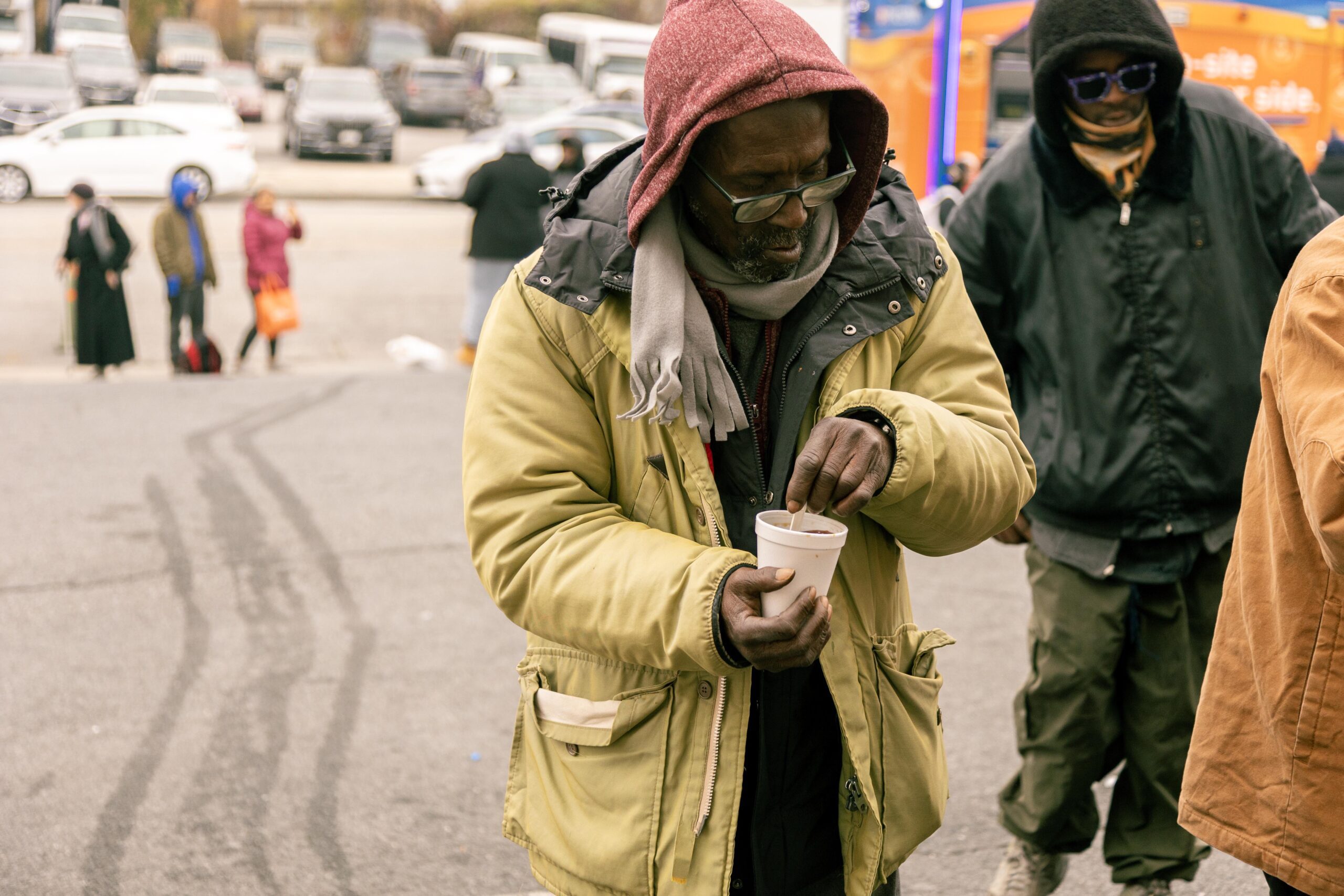 A man in a yellow jacket and red hoodie stirs a cup while standing in a crowded outdoor area, with other people in the background. The scene captures a moment of community interaction during a chilly day.