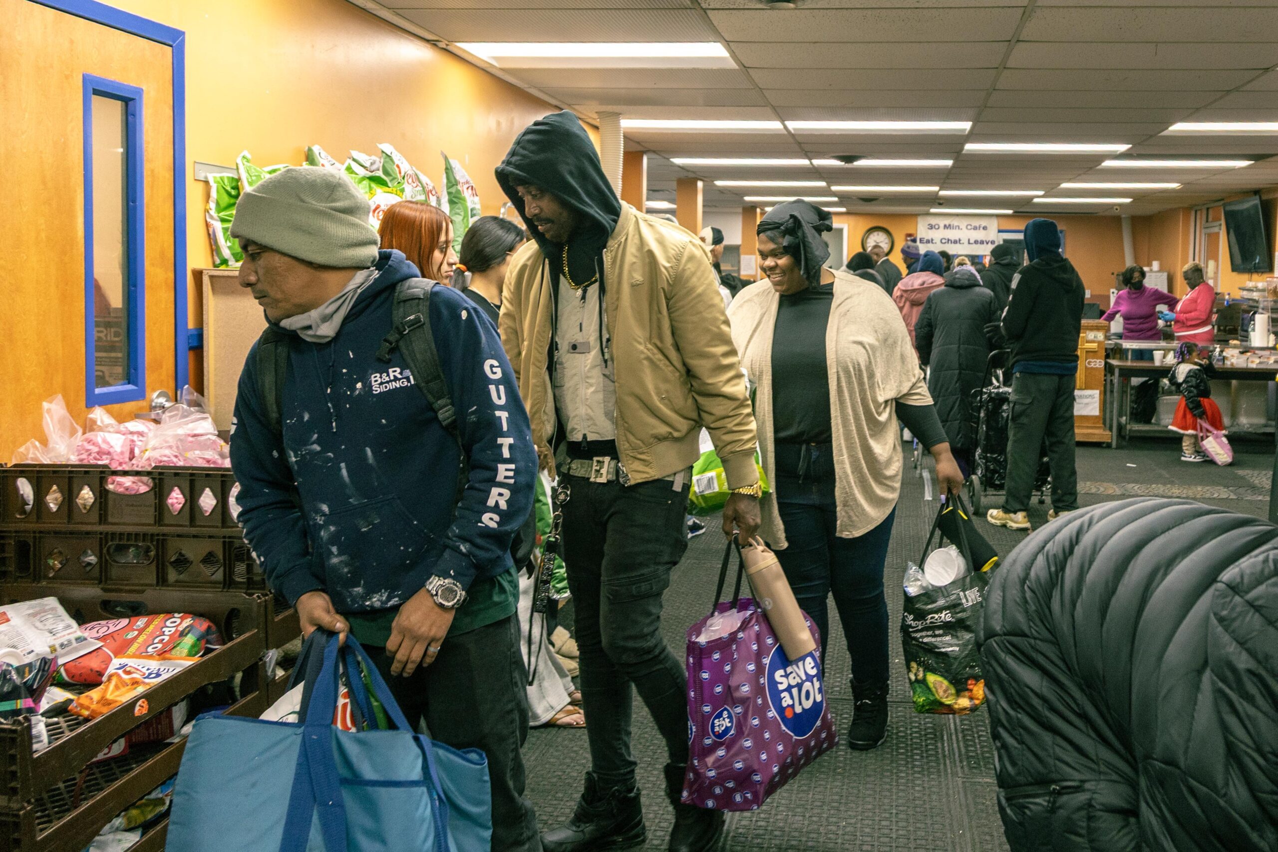 People participating in a community food distribution event, carrying bags filled with groceries, inside a brightly lit room with shelves of food items.