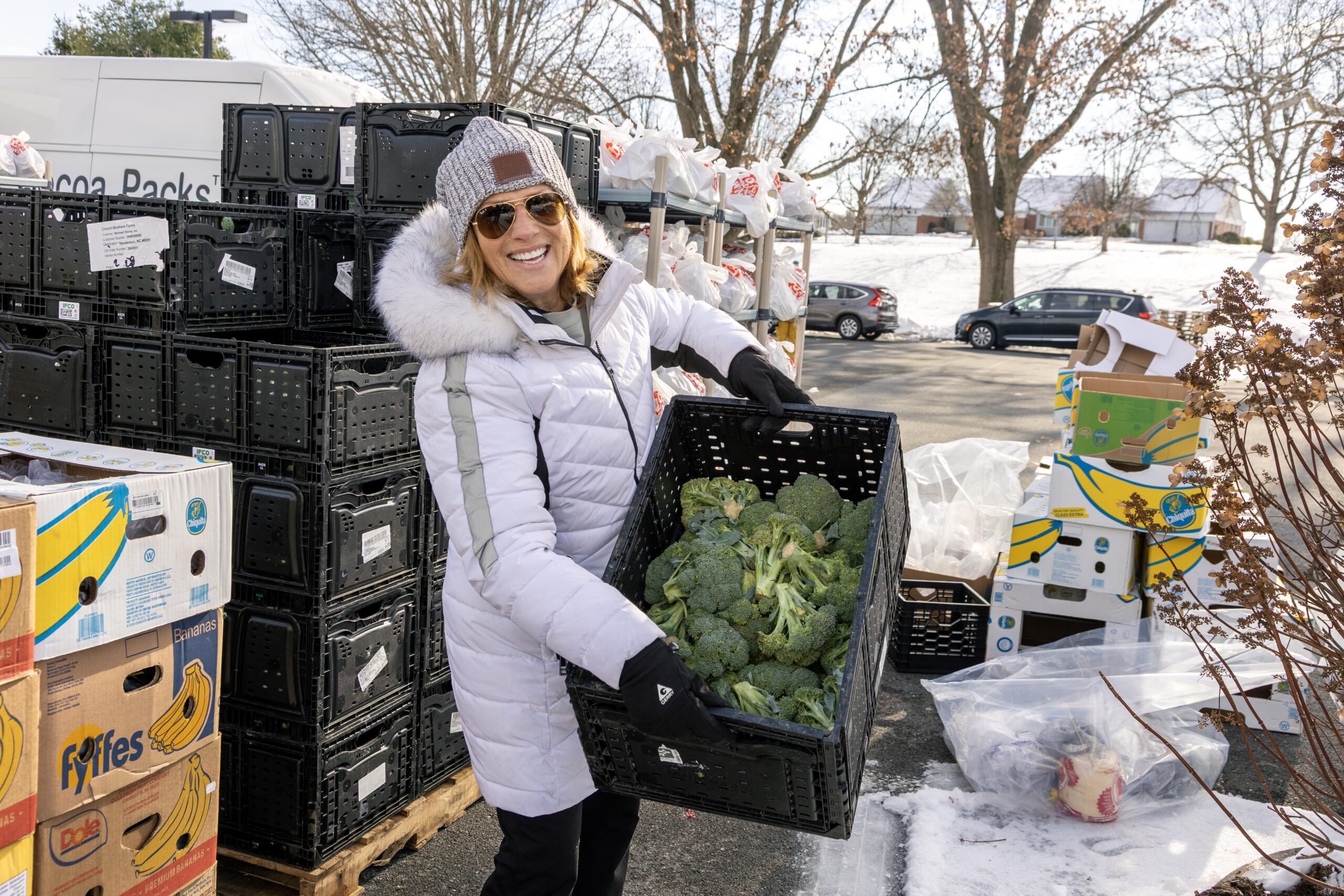 Woman wearing a winter coat and hat joyfully carries a crate of fresh broccoli at a food distribution site, surrounded by stacked crates and bags of produce in a snowy outdoor setting.