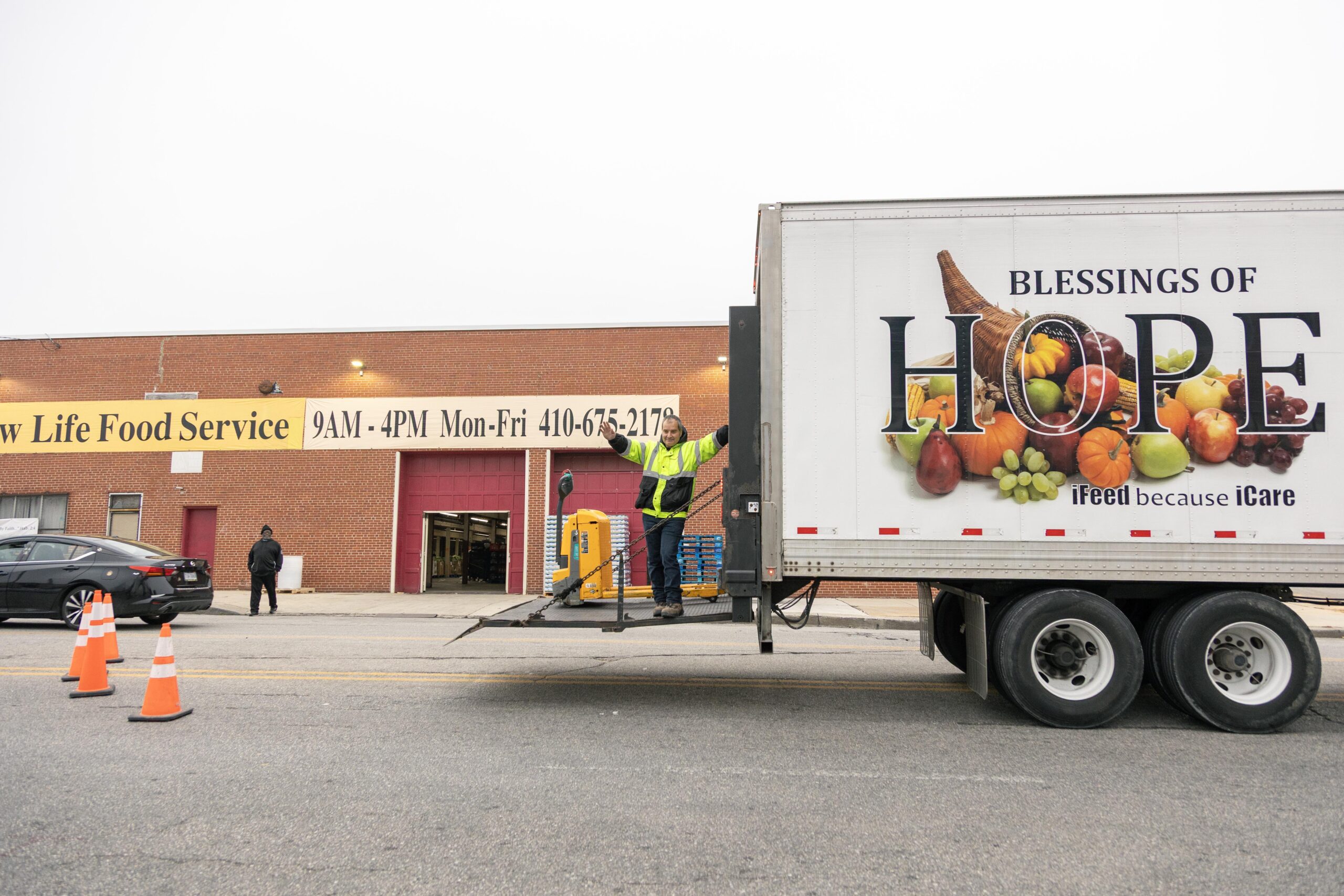Delivery truck at New Life Food Service, showcasing the "Blessings of Hope" message with fresh produce. A worker in a reflective vest stands on the truck's lift, with a brick building displaying operational hours and contact information in the background.