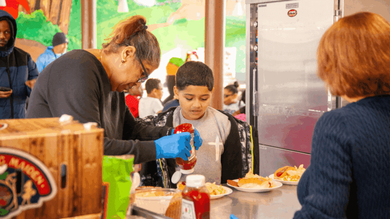 A volunteer serves food to a young boy at a community event, as he adds ketchup to his plate. The scene captures the warmth of community support and the joy of sharing meals.