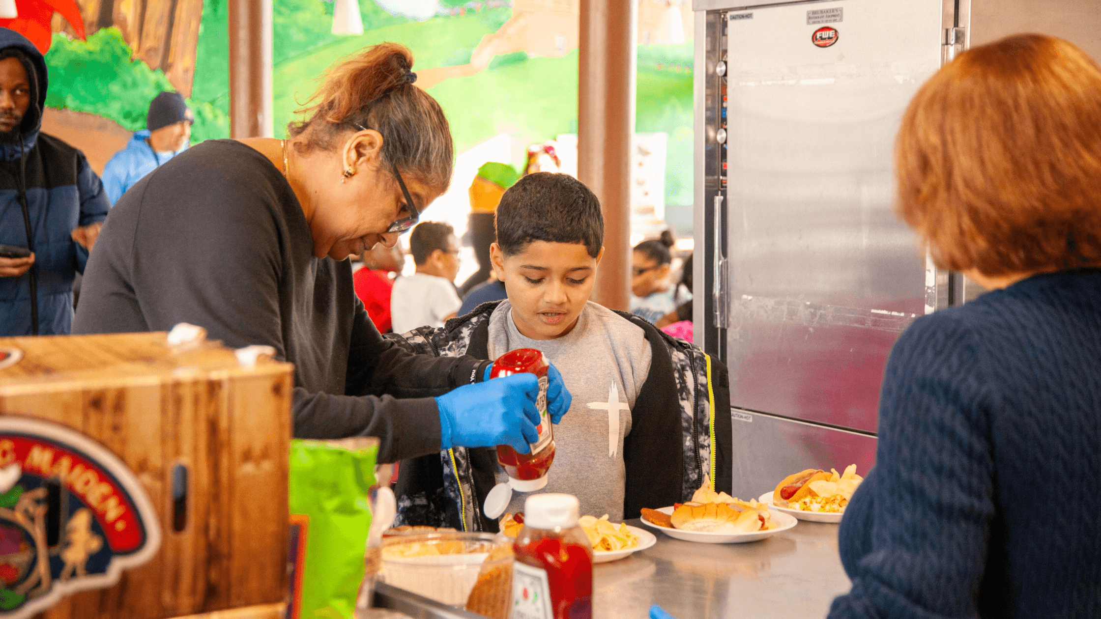 A volunteer serves food to a young boy at a community event, as he adds ketchup to his plate. The scene captures the warmth of community support and the joy and faith of sharing meals.