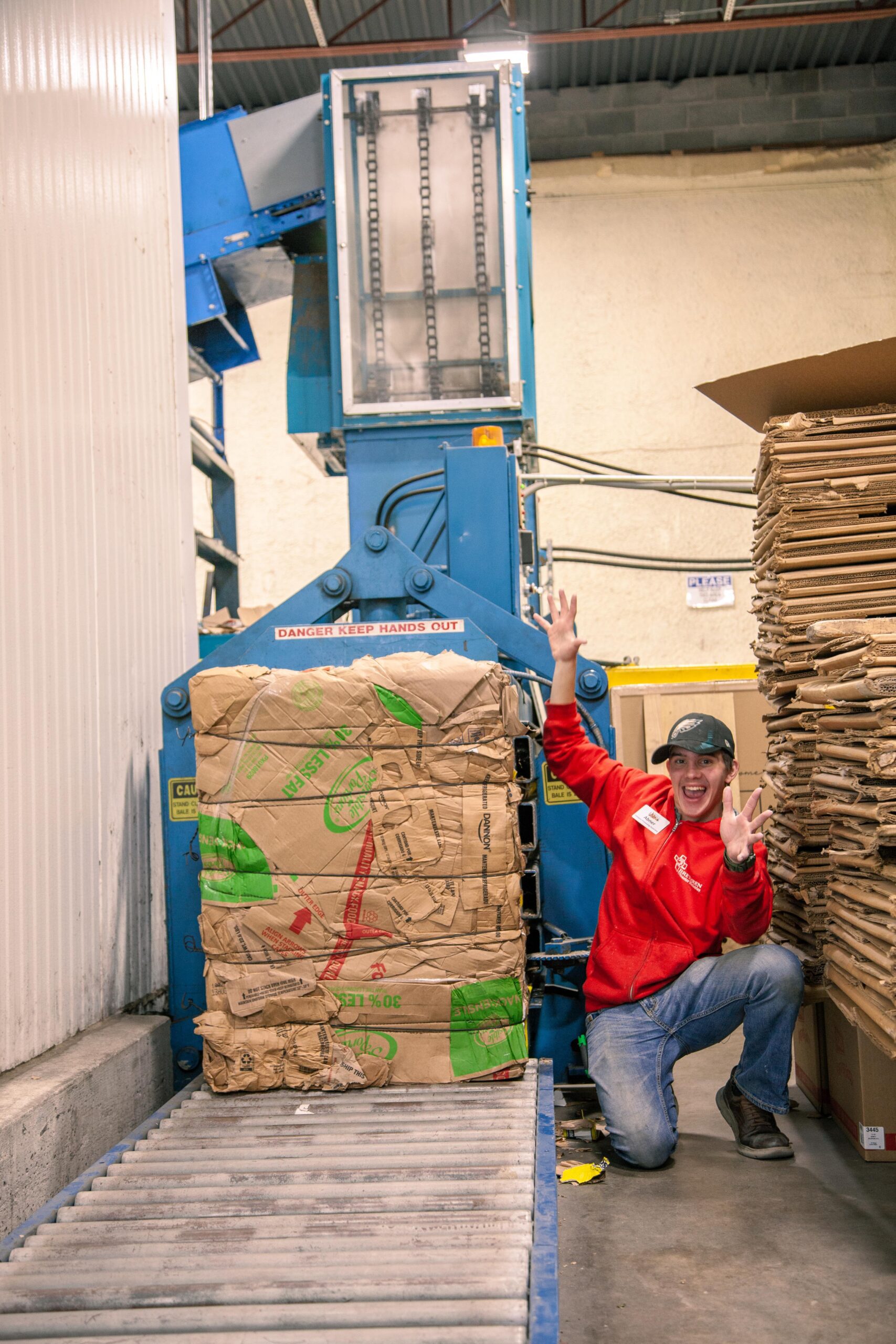 Employee posing next to a cardboard baler in a recycling facility, with stacked cardboard boxes on a conveyor belt and safety signs visible in the background.