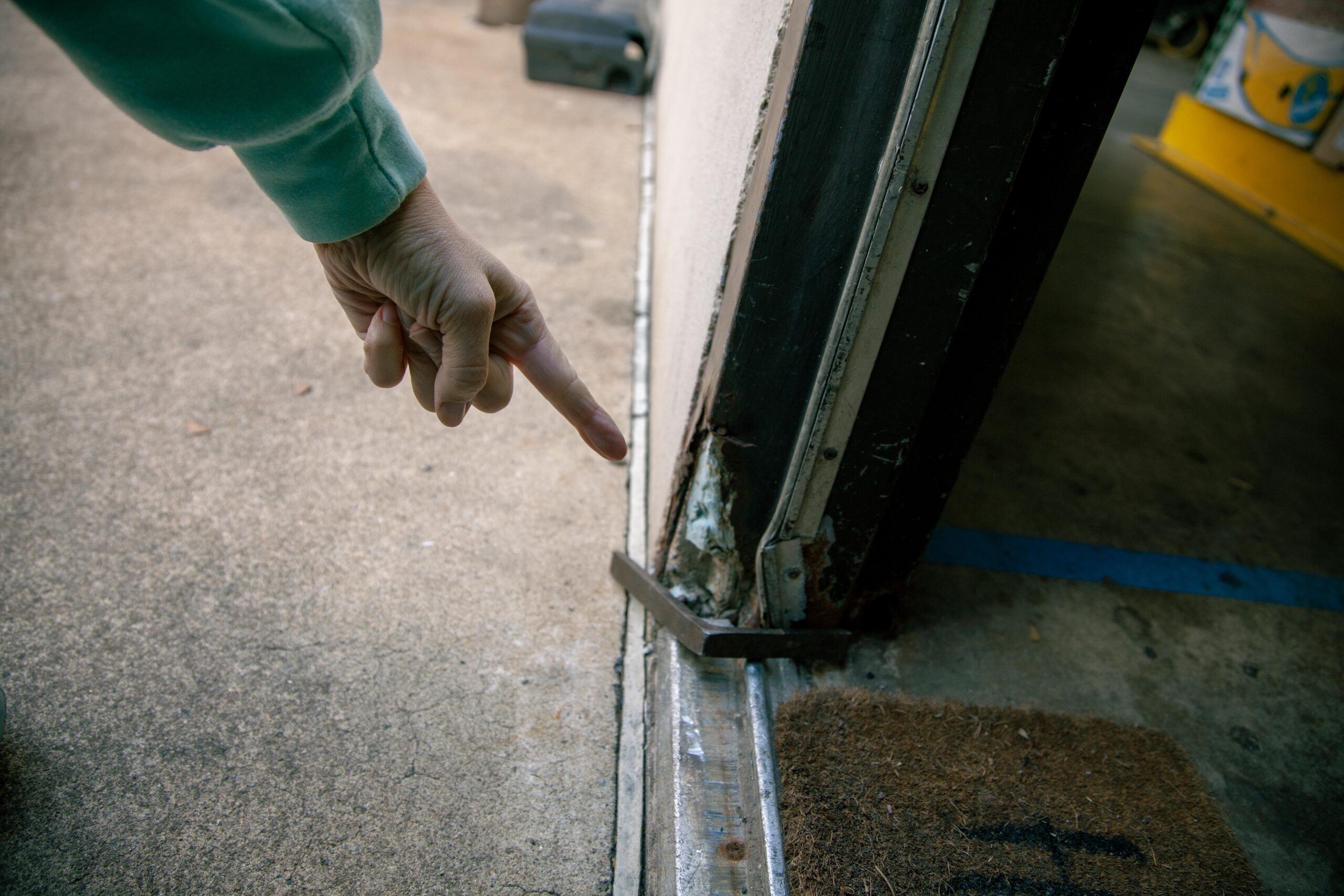 A hand pointing at the bottom corner of a garage door, highlighting rust and damage at the door's seal against the concrete floor. A doormat is visible nearby, suggesting wear and potential water intrusion issues.