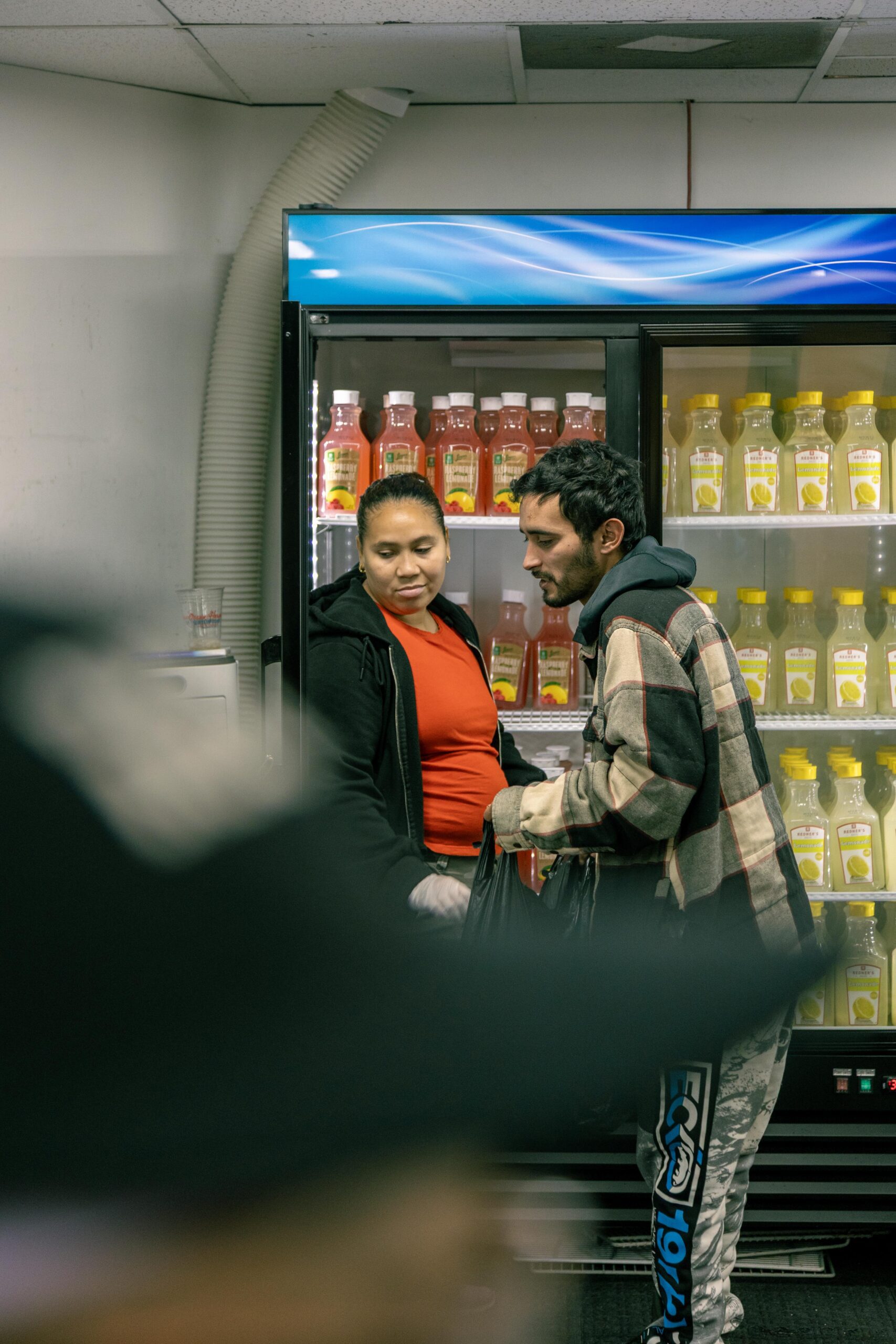 A woman in a black hoodie and red shirt interacts with a man wearing a plaid jacket in front of a refrigerated display filled with beverages, including lemonades and fruit drinks. The setting appears to be a convenience store or market.