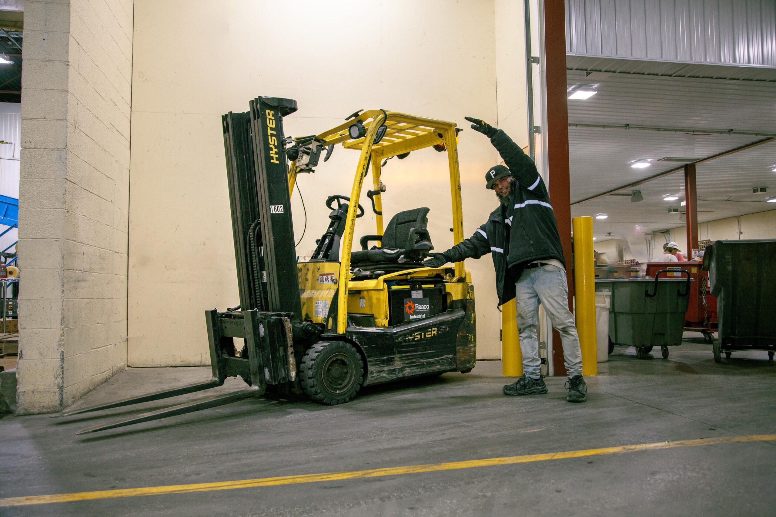 Warehouse worker proudly showcasing a yellow Hyster forklift in a busy industrial setting, highlighting its features and functionality.