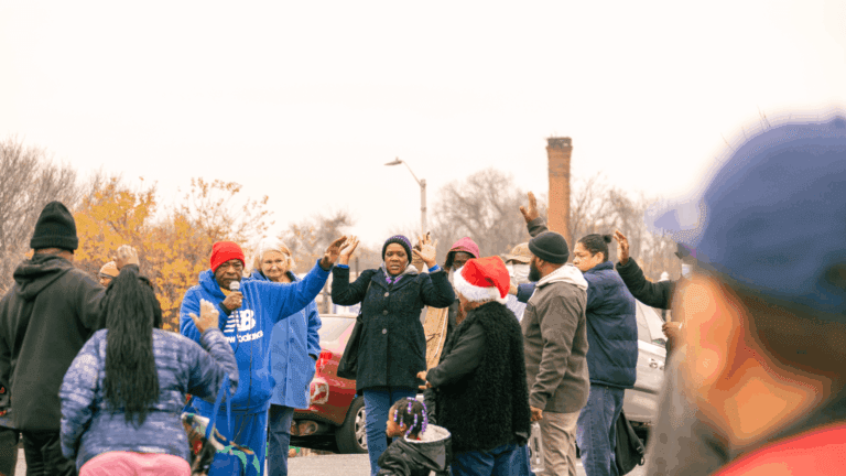 Community members gather in a parking lot, engaged in a lively discussion with raised hands. A speaker in a red hat addresses the crowd, while others listen attentively. The scene reflects community activism and unity during an outdoor event in an urban setting.