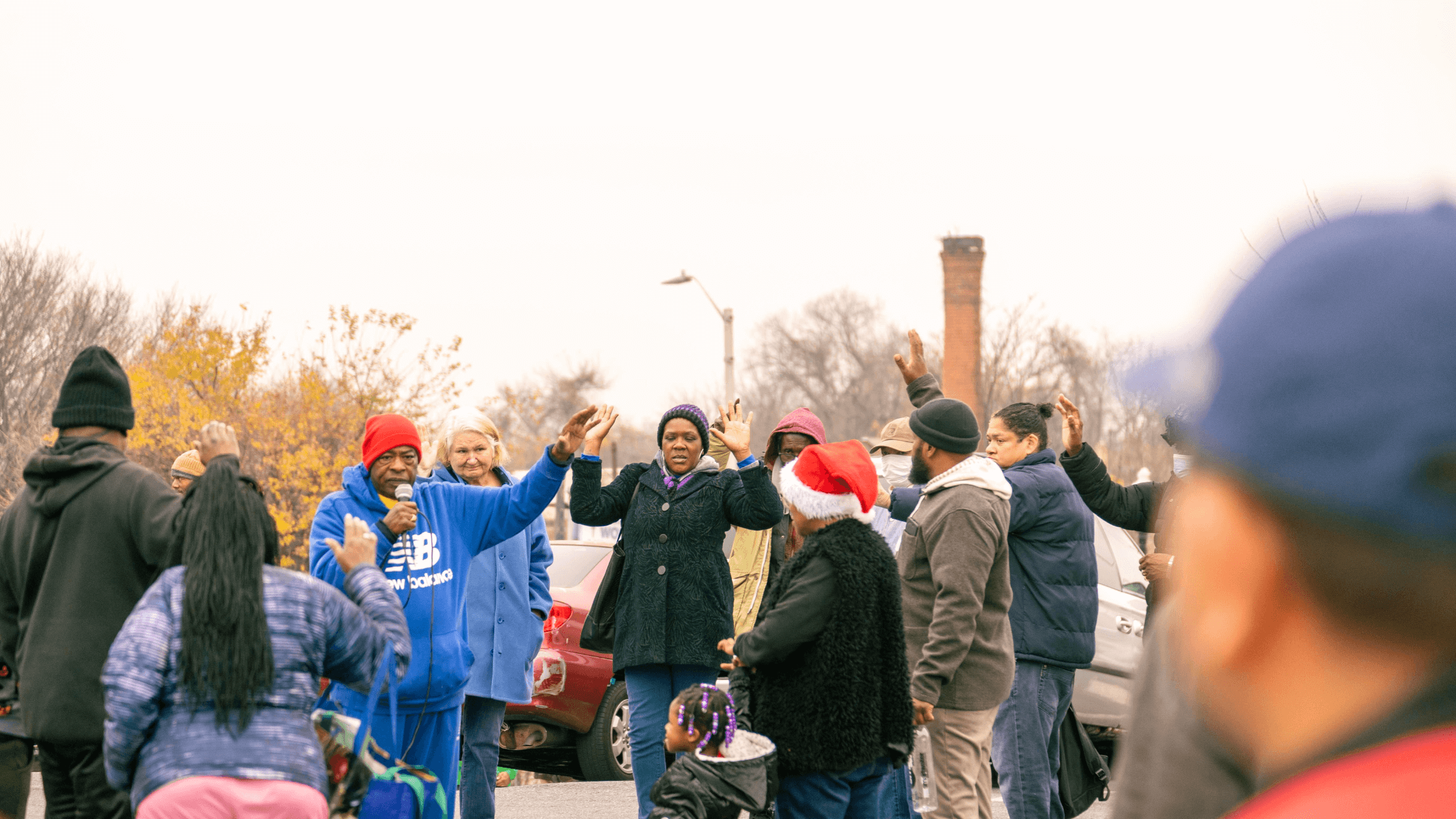 Community members gather in a parking lot, engaged in a lively discussion with raised hands. A speaker in a red hat addresses the crowd, while others listen attentively. The scene reflects community activism and unity during an outdoor event in an urban setting.