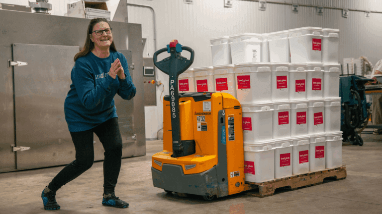 Woman smiling and posing next to an electric pallet jack loaded with white containers labeled "food.gives" in a warehouse setting, highlighting food distribution efforts.