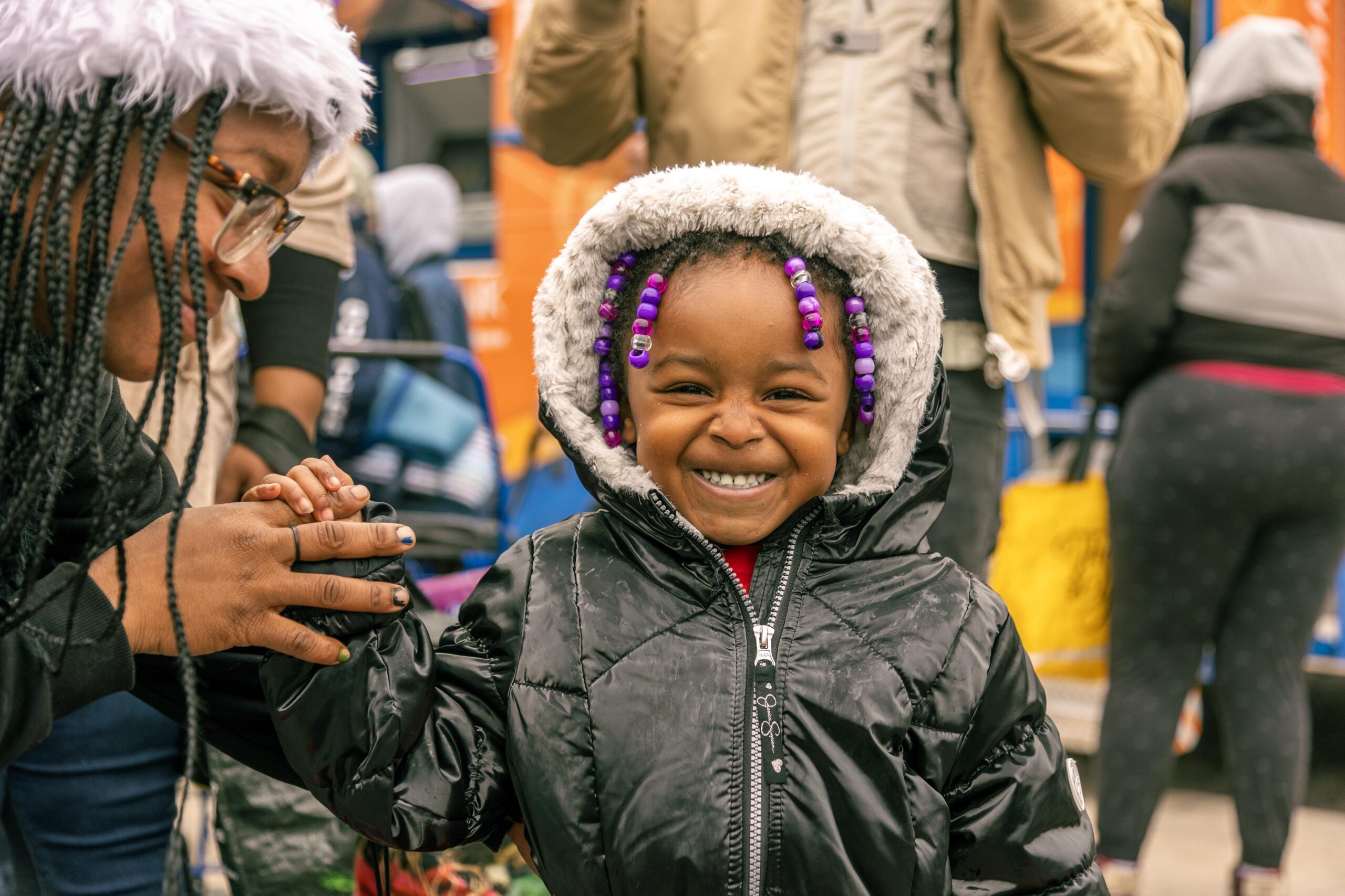 Smiling young girl in a black winter jacket with a furry hood, wearing colorful beaded braids, holding hands with an adult in a winter setting, surrounded by other people and colorful backgrounds.