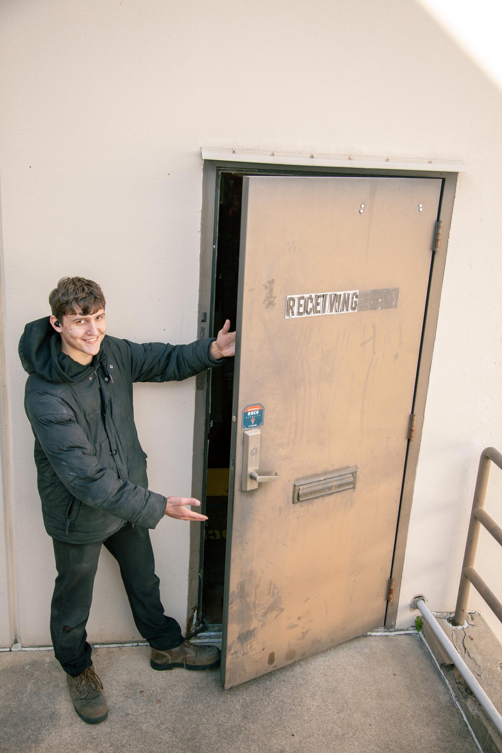 Young man in a black jacket standing beside an open receiving door, gesturing towards it with a smile. The door is labeled "RECEIVING" and is located in a well-lit outdoor area.