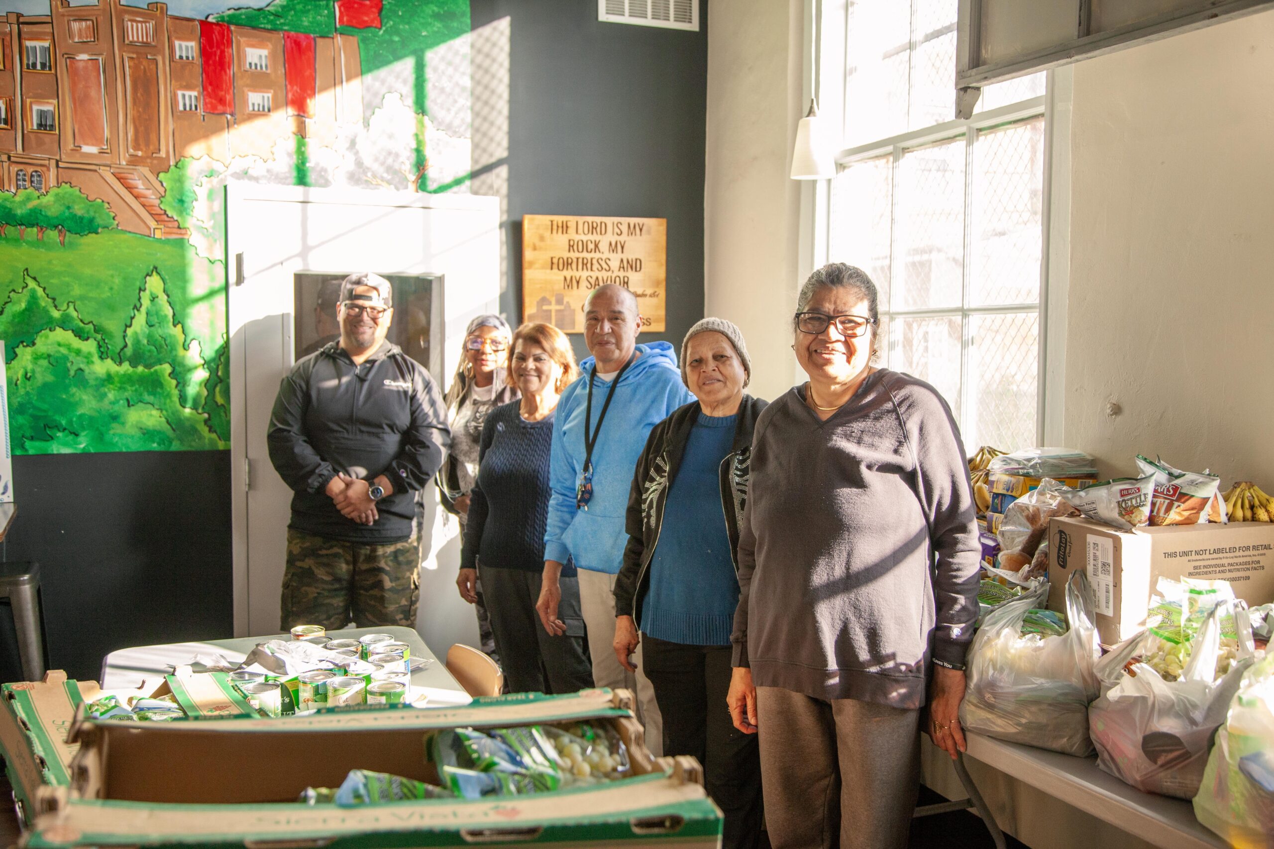 Group of volunteers standing in a community center, surrounded by food donations, with a mural in the background depicting greenery and buildings. The setting highlights community support and collaboration in addressing food insecurity.