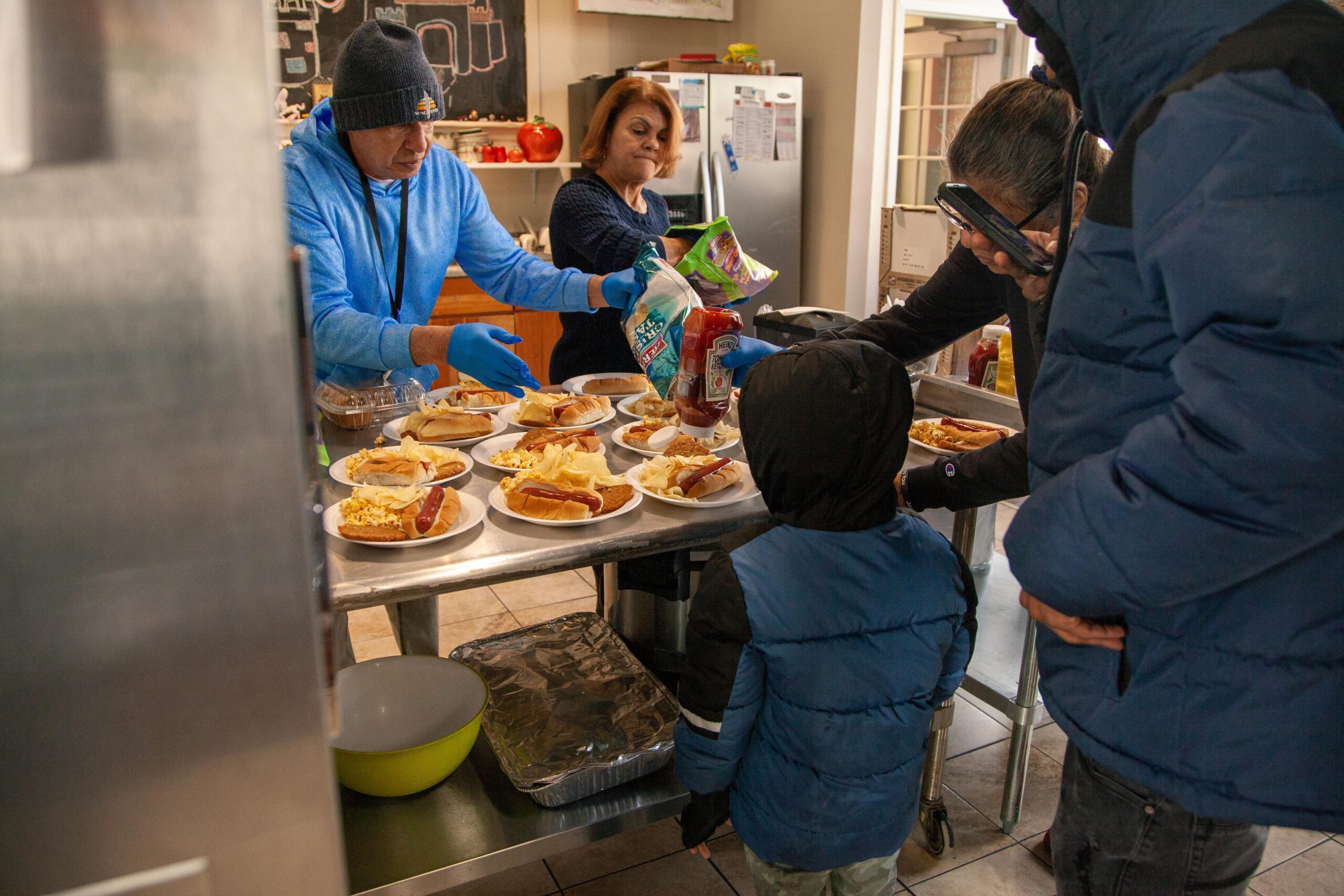 Volunteers serving hot dogs and snacks at a community meal event, with a child watching as food is prepared. The setting includes a kitchen with plates of food and condiments, capturing a moment of generosity and community engagement.