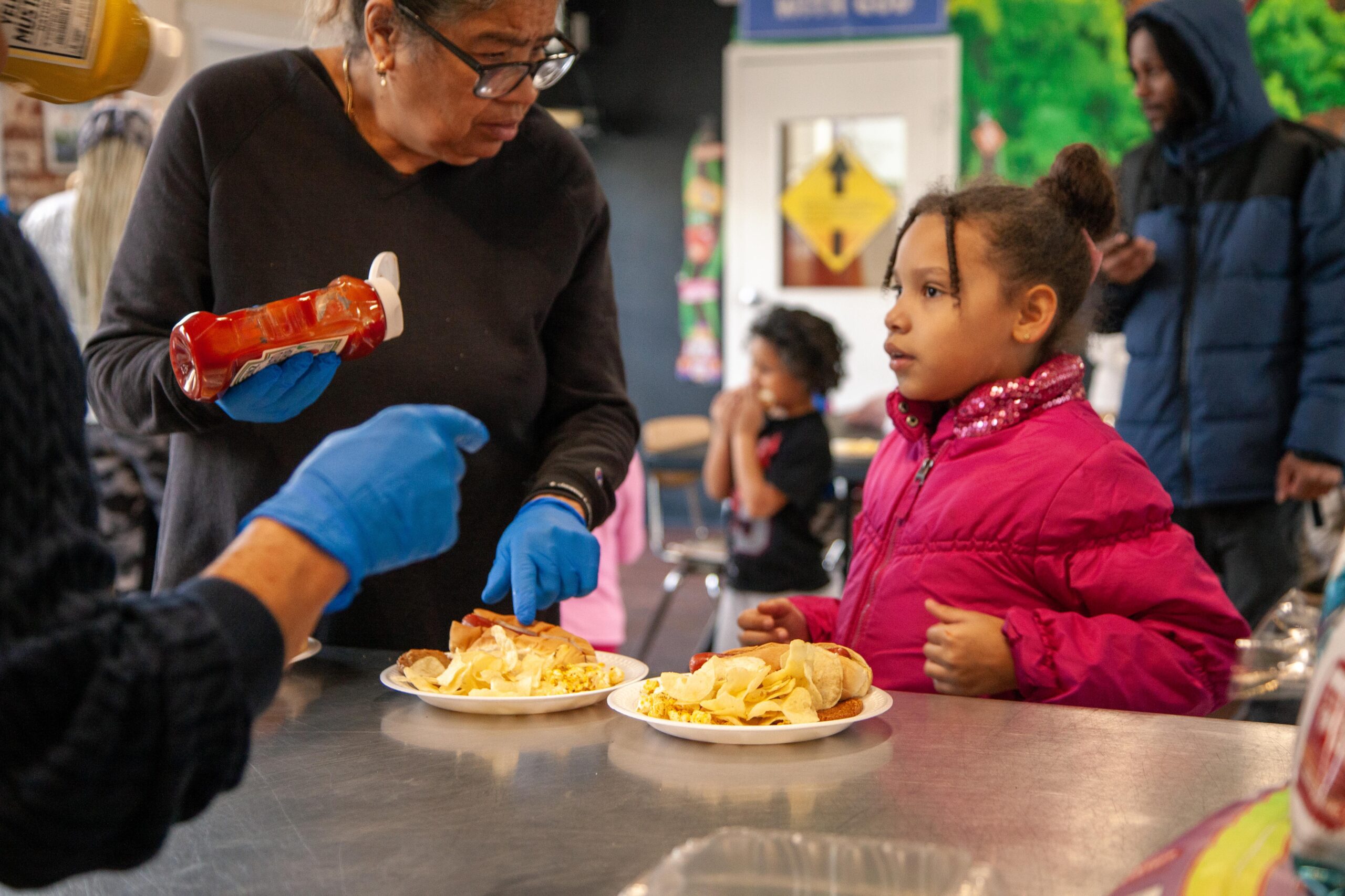 Woman serving food to a young girl at a community event, with plates of hot dogs and chips on the table. The woman is holding a bottle of ketchup, wearing blue gloves, while the girl, dressed in a pink jacket, looks on attentively. Background features other children and a colorful mural.