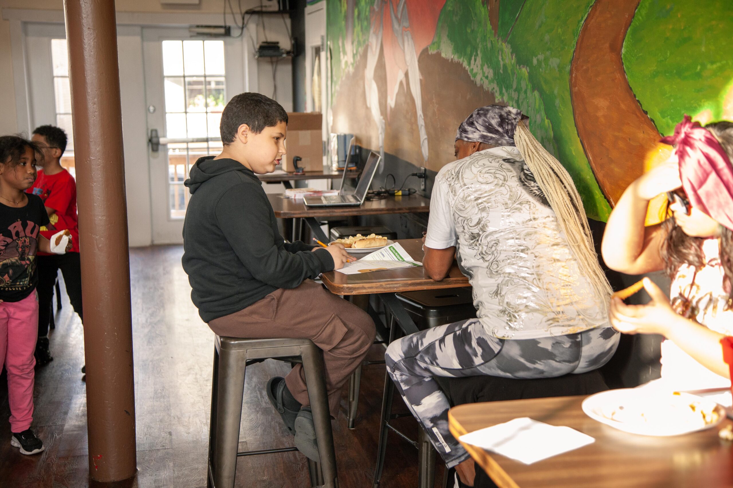 Children engaging in an interactive learning environment at a community center, with one boy sitting at a table working on a project while others socialize nearby. The colorful mural in the background adds a vibrant touch to the space.