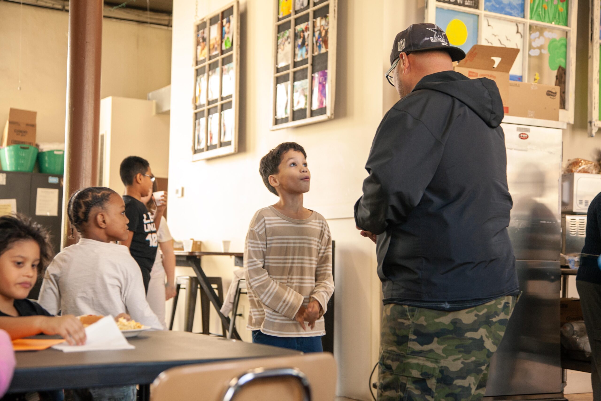 Children interacting in a community center, with a young boy in a striped shirt engaging in conversation with an adult. The background features artwork displayed on the walls and children enjoying snacks at a table.