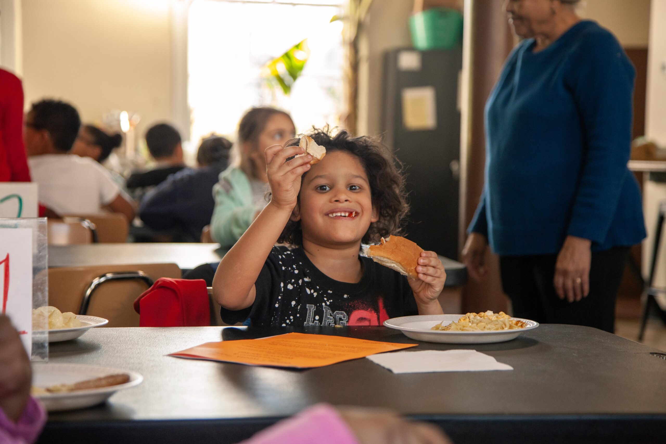 Smiling child enjoying a meal in a communal dining setting, holding a piece of bread and sitting at a table with plates of food and colorful papers around. Background features other children and an adult, creating a warm and inviting atmosphere.