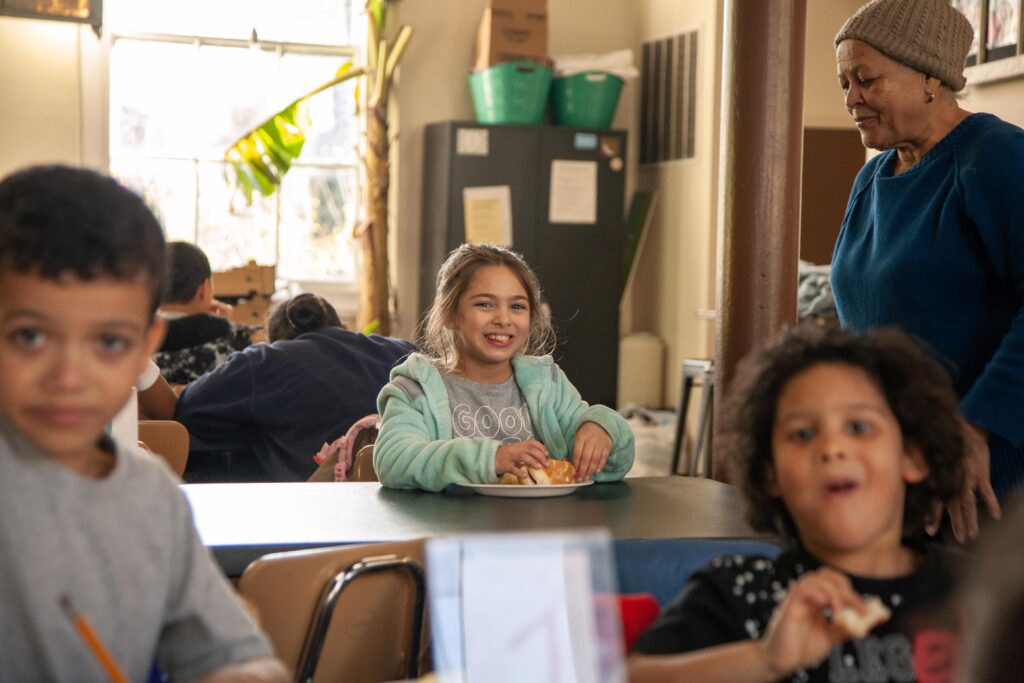 Children enjoying a meal in a communal setting, with a smiling girl in a turquoise sweater holding a plate of food, while an older woman observes nearby. The background features other children seated at tables, creating a warm and engaging atmosphere.