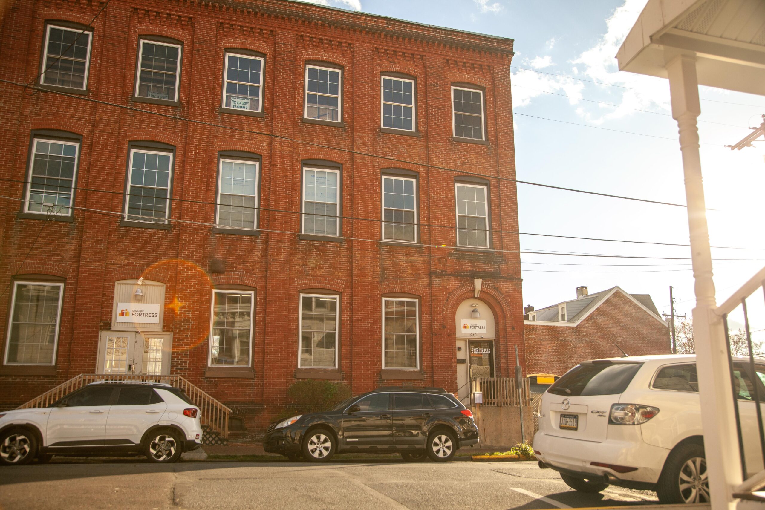 Historic brick building with large windows, featuring the Fortress logo above the entrance; parked cars are visible in front, with a sunny sky in the background.