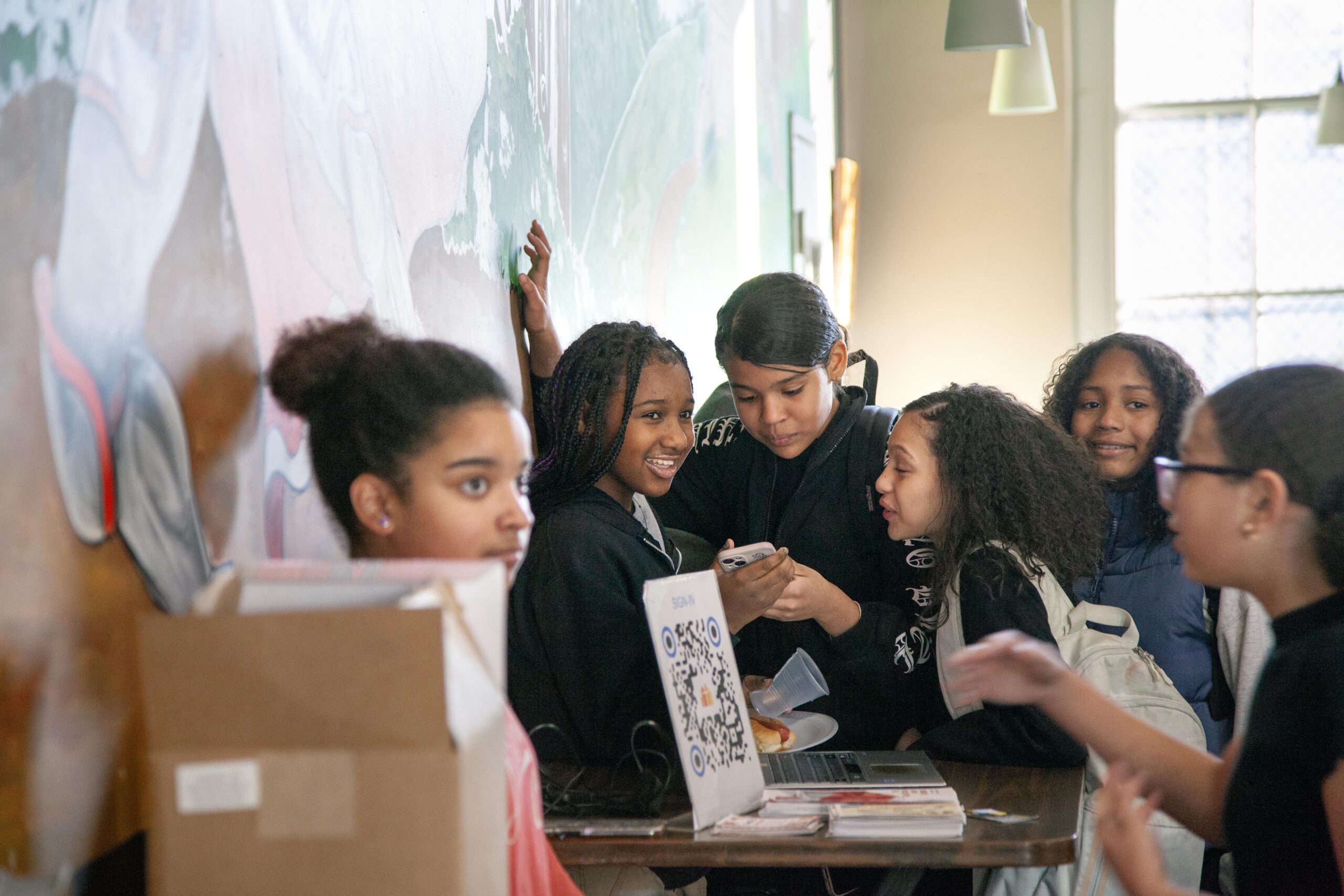 Group of diverse teenagers engaging and laughing together in a vibrant café setting, with one girl showing something on her phone. A laptop and a plate of food are visible on the table, alongside a QR code sign. The background features a colorful mural, creating a lively atmosphere.